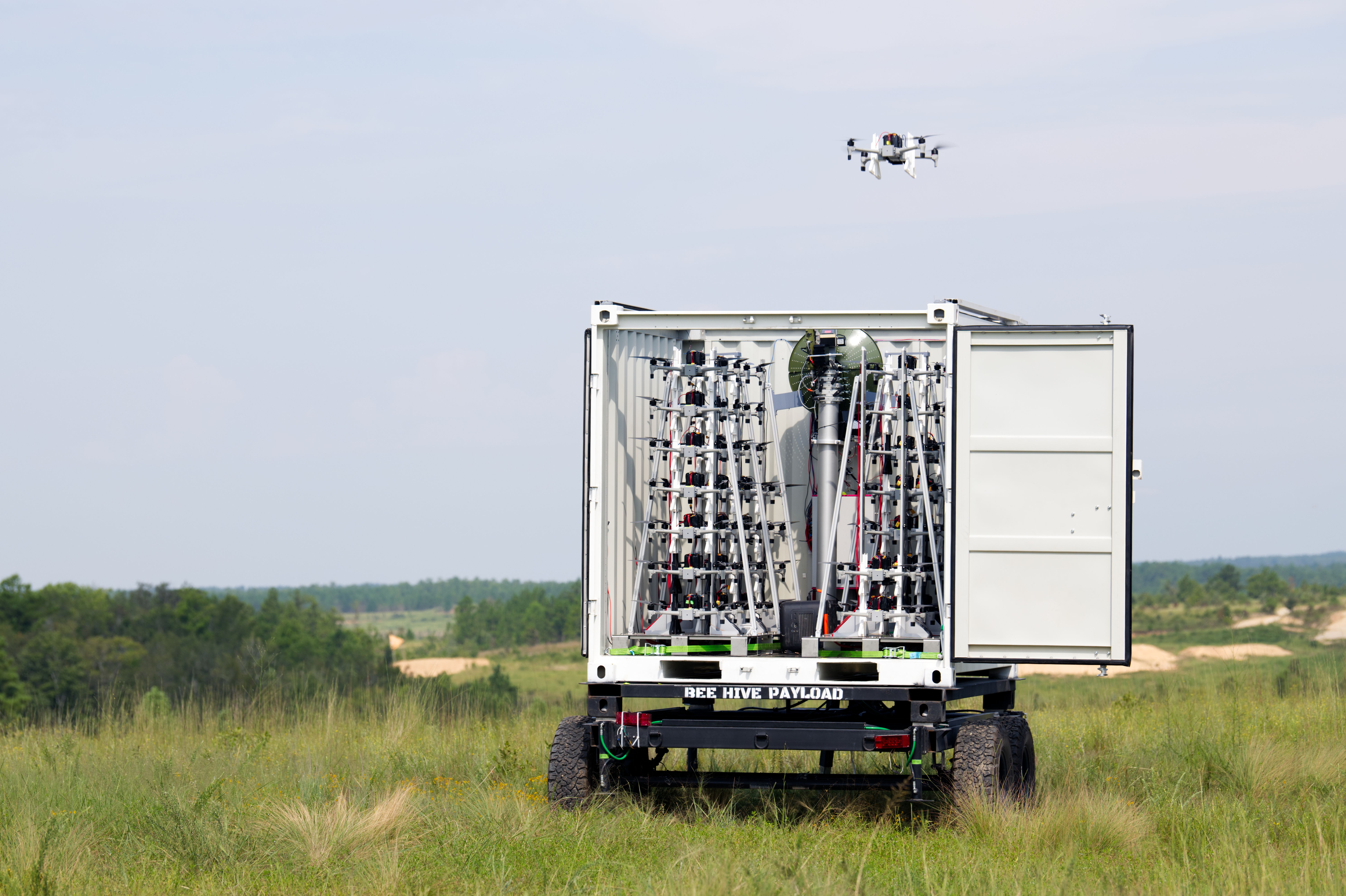 A white shipping container on a trailer opens to reveal a dense rig of drones and wiring. A small quadcopter drone hovers in the pale blue sky above a grassy field.