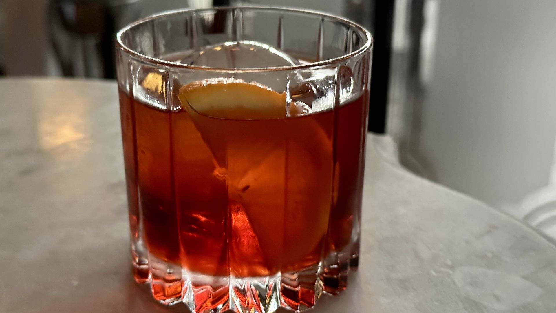 A cocktail glass filled with a ruby red liquid, spherical ice and a lemon wedge.