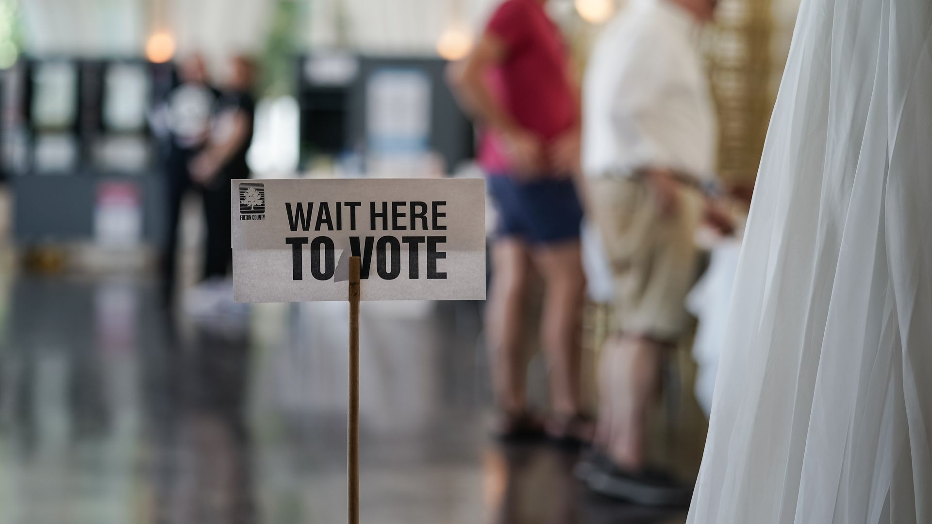 A sign reads "wait here to vote" as some people are seen in the background