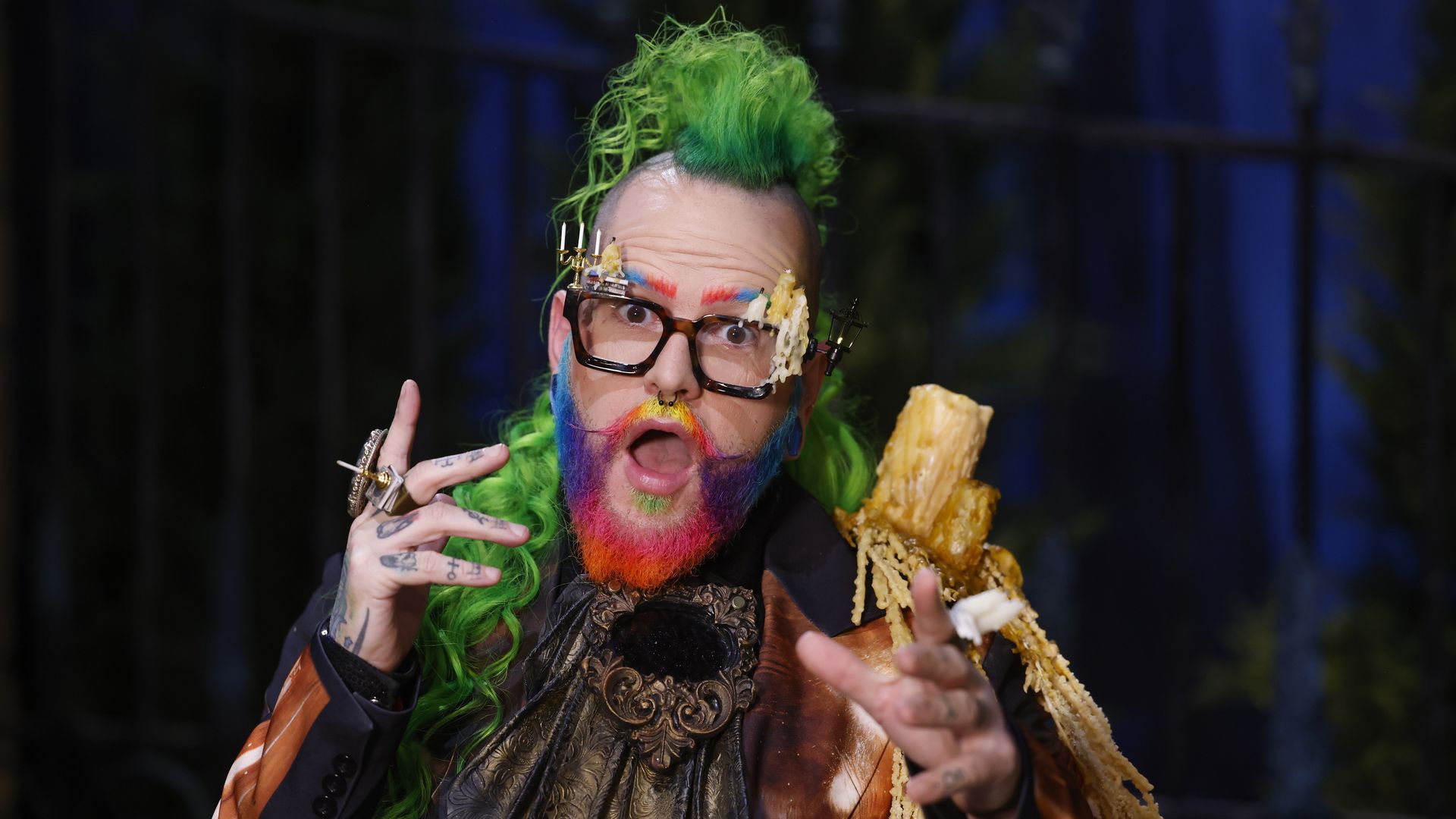 Andrew Fuller with a bright green mohawk, rainbow face paint, and oversized decorative glasses, holding a pastry in a dark blue, blurred background.