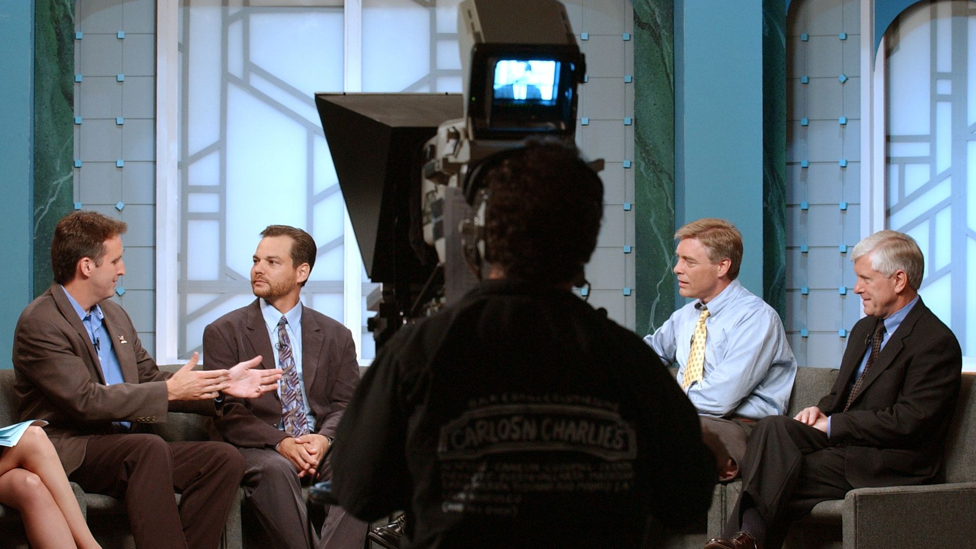 Four men in suits and a woman with beige heels sit on a TV talk show set with a cameraman filming them. The background is light blue with geometric patterns.