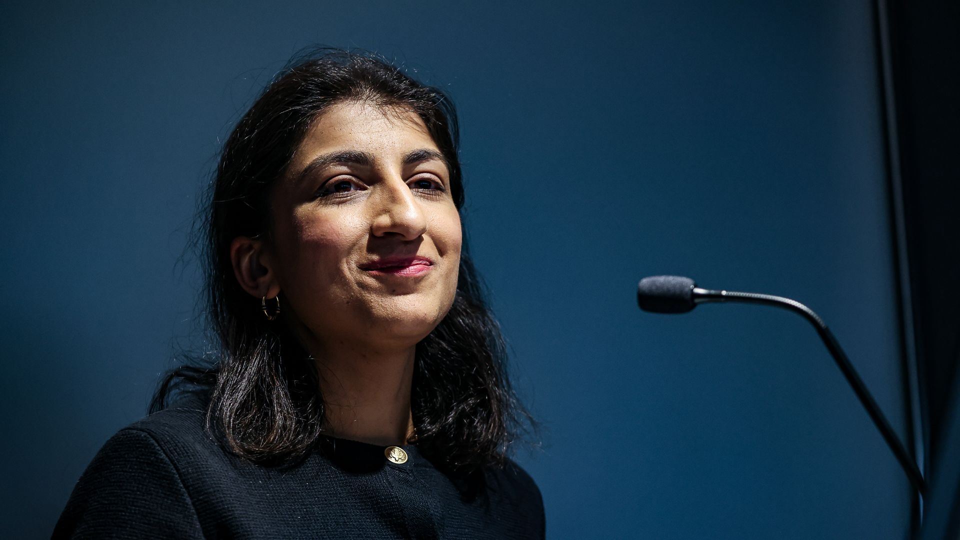 FTC chair Lina Khan speaking at a lectern with a blue backdrop