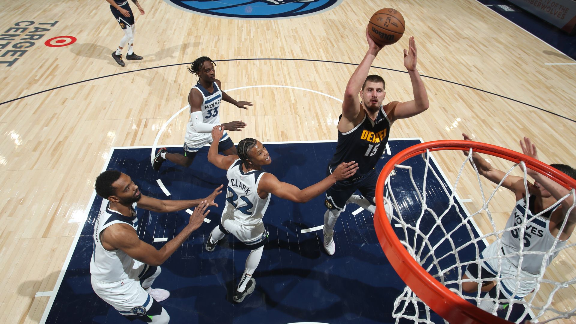 A man in a dark blue jersey drives up to throw a basketball into a hoop while surrounded by four players in white jerseys.