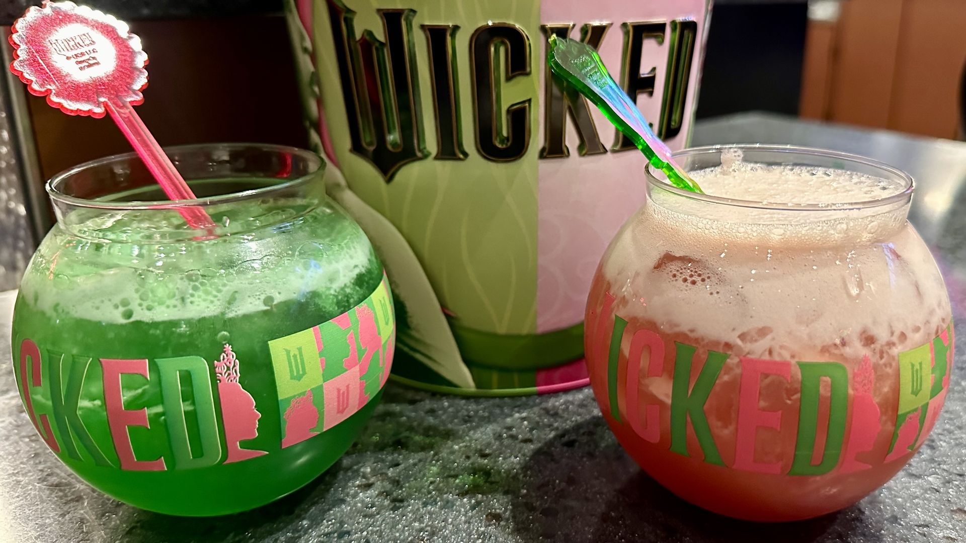 A photo of a green cocktail in a bubble glass and a pink cocktail also in a bubble glass in front of a tin of popcorn with the "Wicked" logo on it