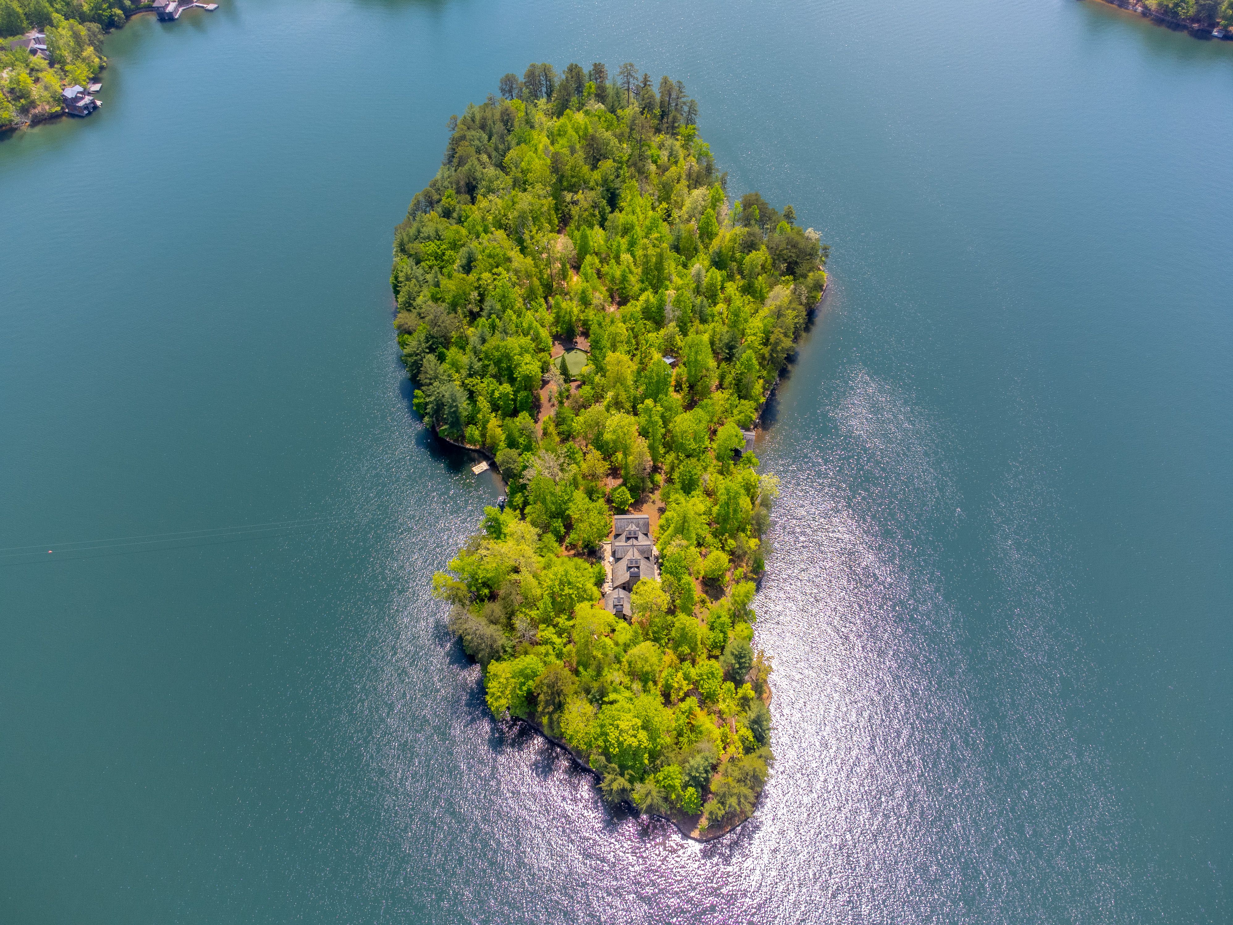 Aerial view of a densely wooded island in a large body of water with a large house visible near the center, sunlight reflecting on the water's surface around the island.