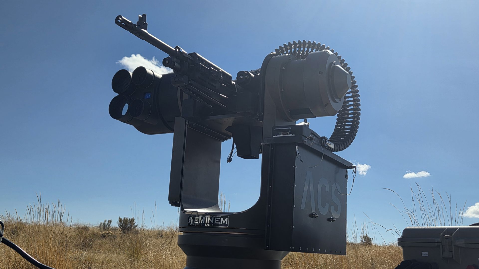 Black automated weapon system labeled ACS mounted on a vehicle, featuring a long barrel gun and belt-fed ammunition, under a clear blue sky with dry grass in the background.