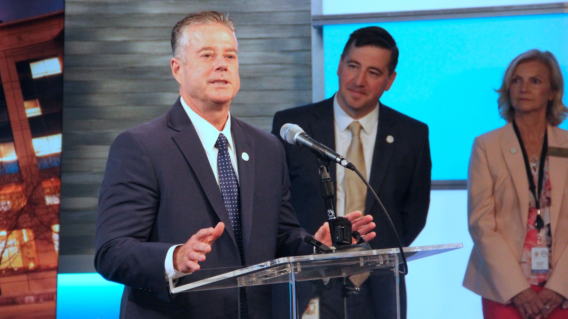 A man in a suit stands in front of a plastic lectern while speaking. 