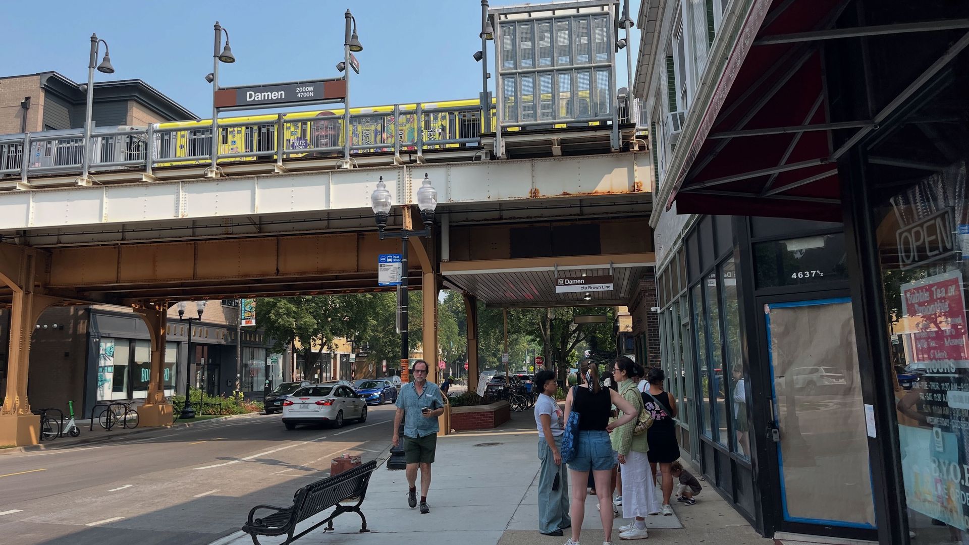 Street scene at Damen CTA Brown Line station with elevated train, people standing on sidewalk near storefronts, and a man walking by a bench under clear blue sky.