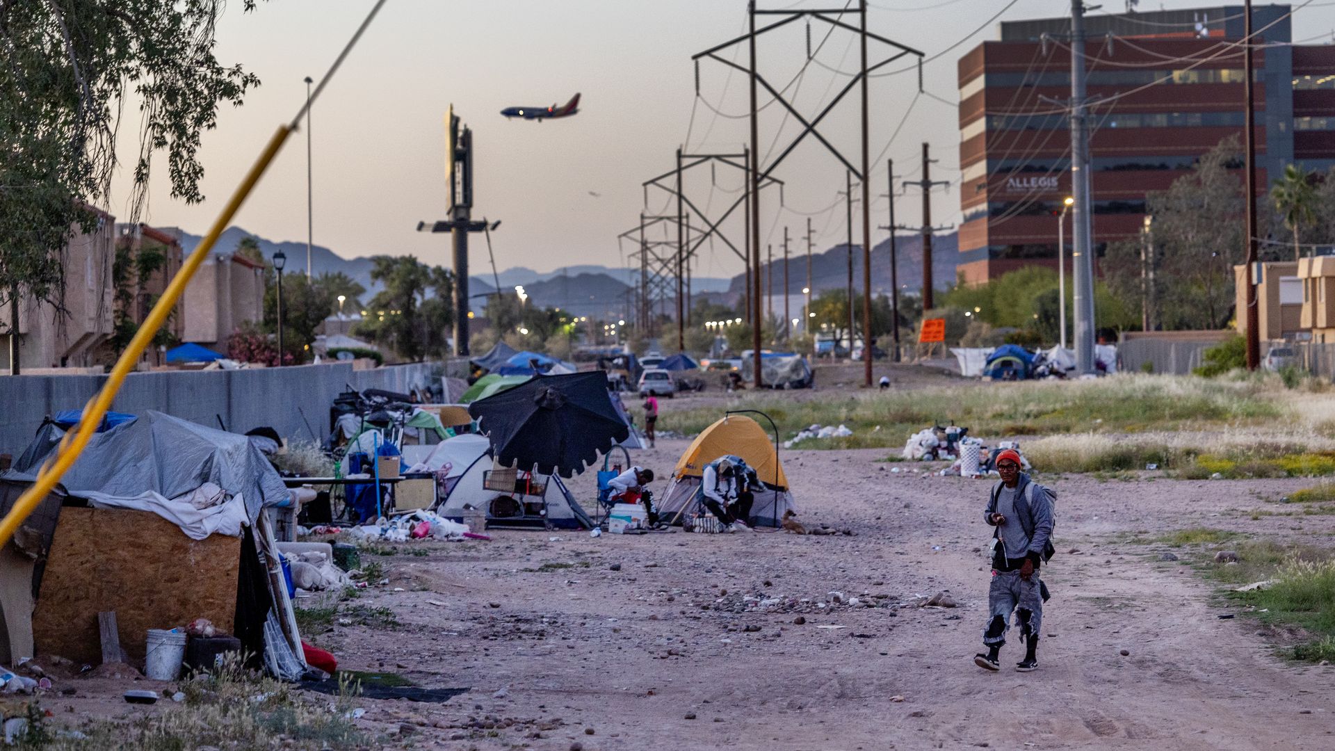 A homeless encampment lines a gray stucco wall on a plot of dirt, with buildings in the background and a disheveled man walking in the foreground. 
