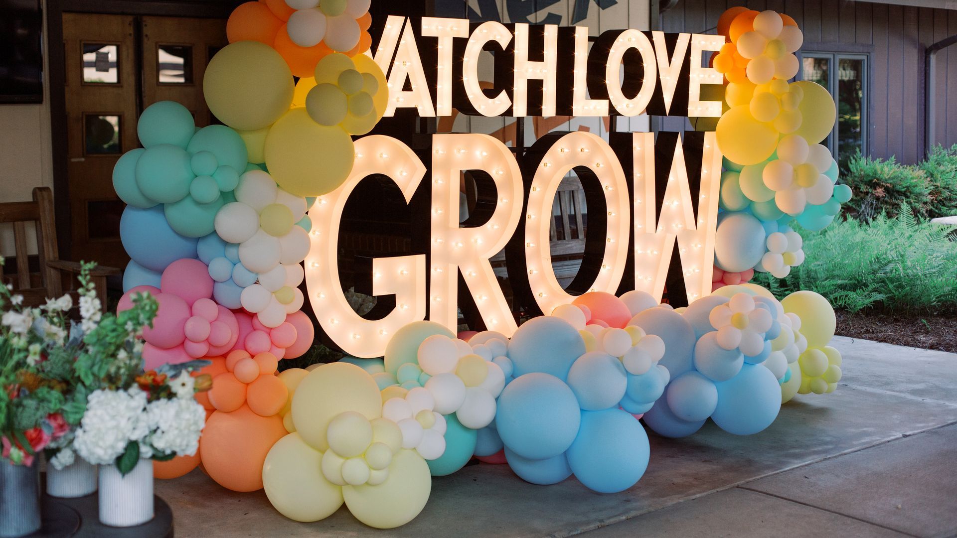 Large illuminated sign reading WATCH LOVE GROW framed by pastel balloon clusters in mint, peach, pink, yellow, and blue, set outside a building with greenery and potted flowers nearby.