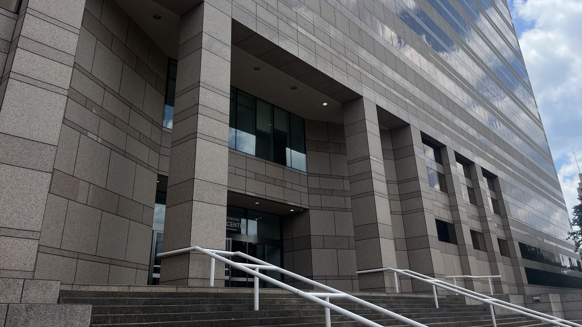 A modern gray stone office building seen at an angle, with reflective glass windows showing clouds and a blue sky. Foreground features steps and white handrails.
