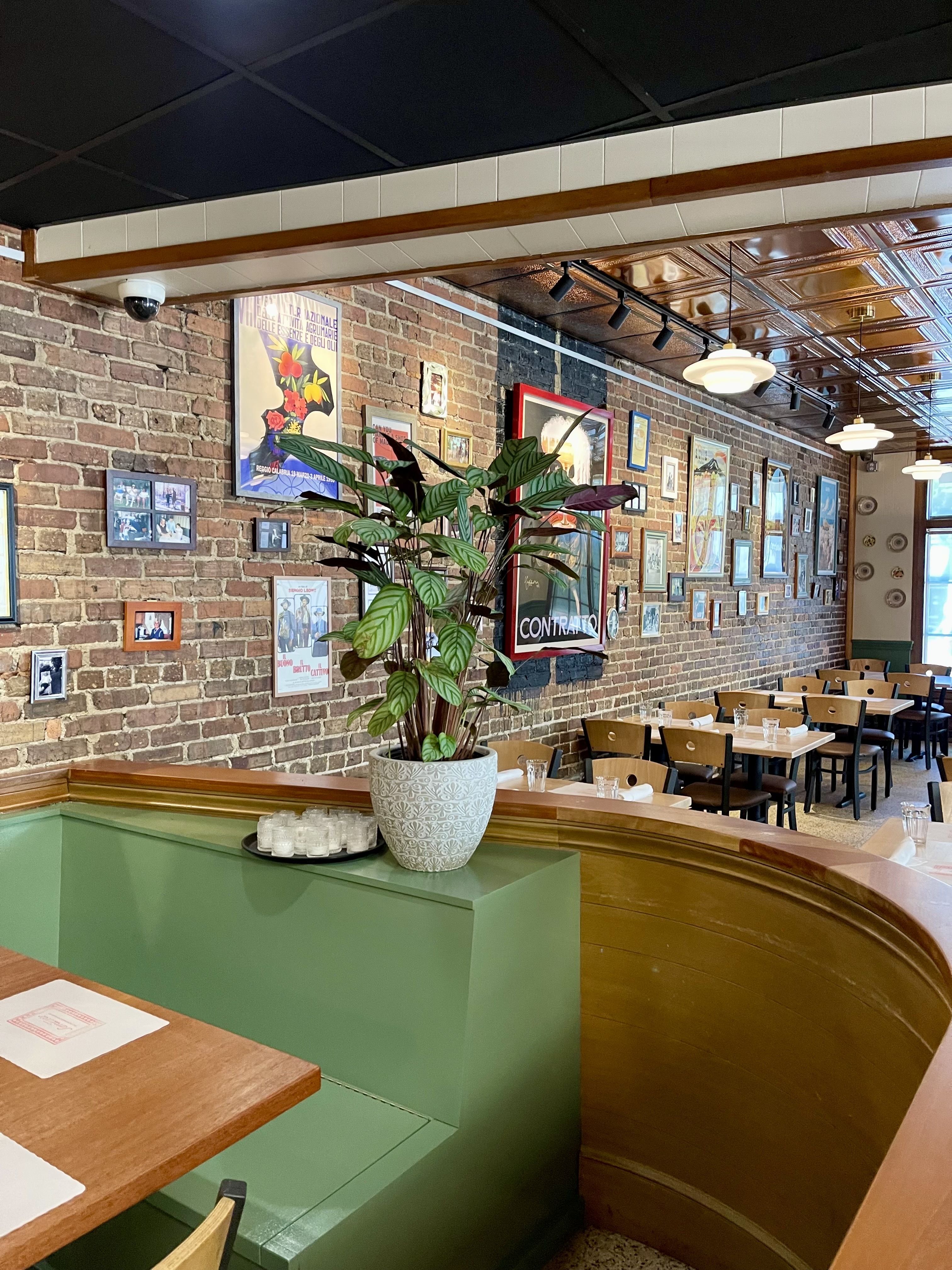 Interior of a restaurant with green booth seating, wooden tables, and chairs. Exposed brick wall adorned with numerous framed pictures and posters. A large potted plant on the booth divider.