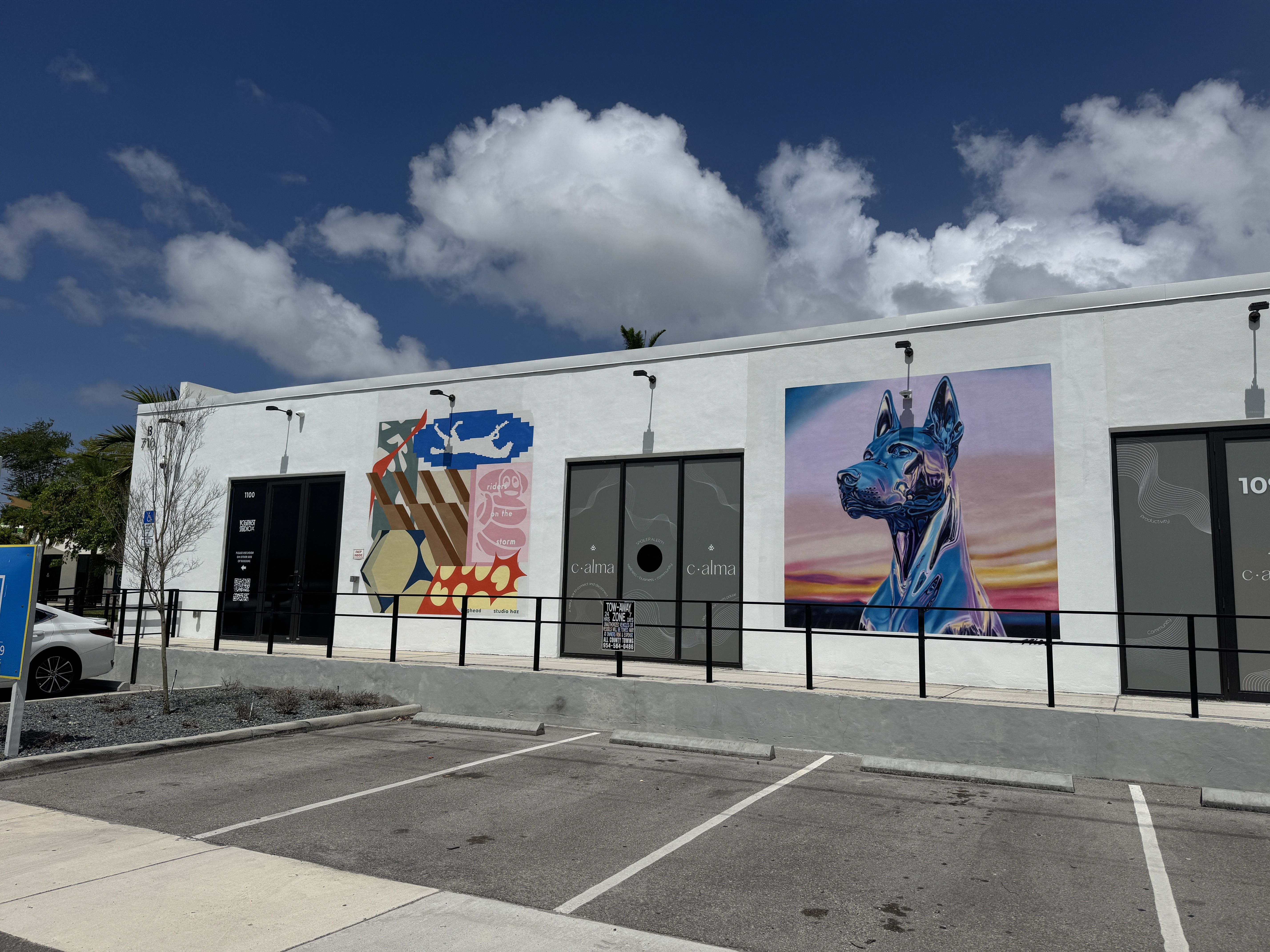 White commercial building with colorful mural panels along the facade and a glossy blue animal artwork on the right, under a blue sky with white clouds; a parking lot in foreground.