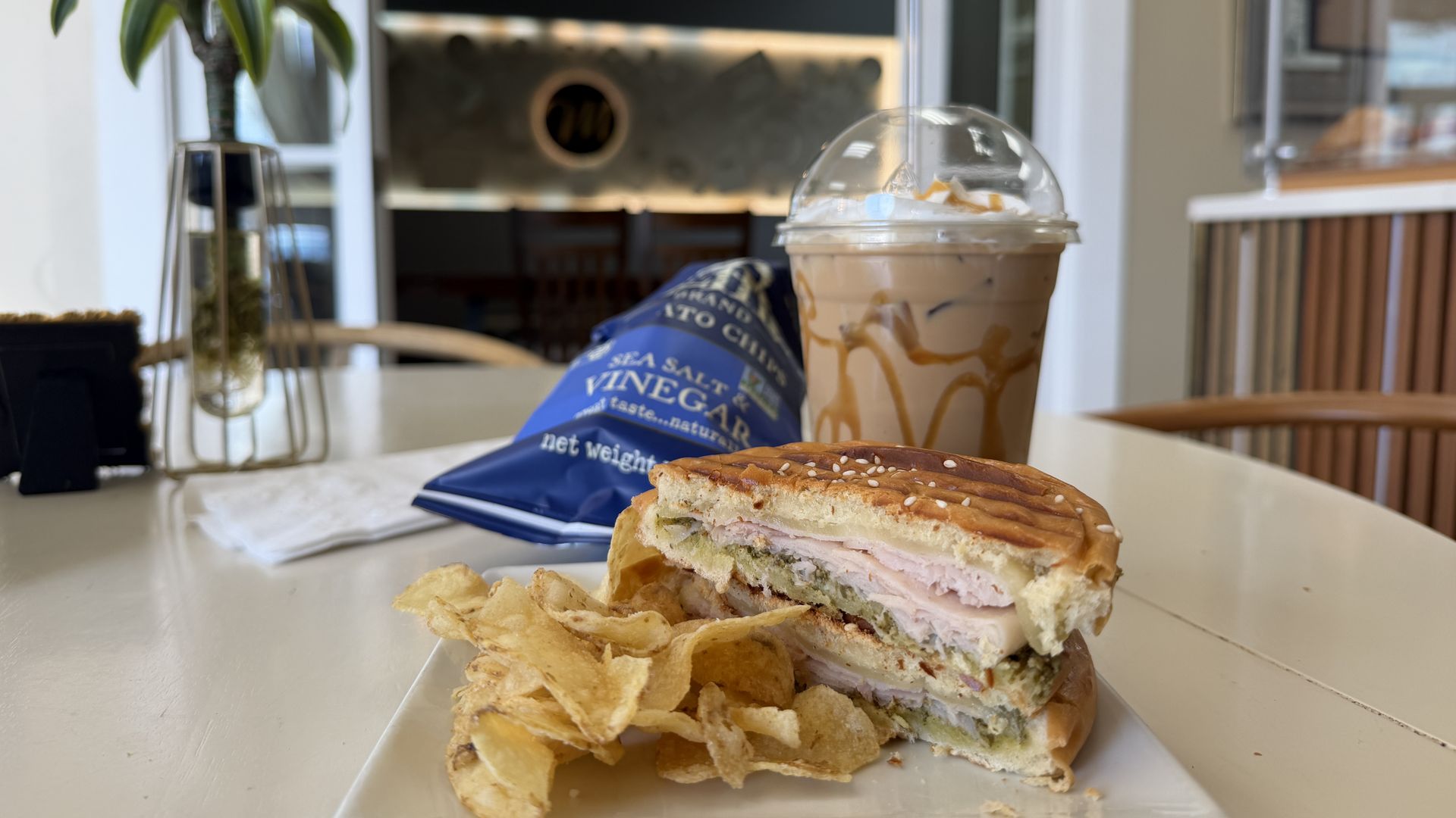 Sesame bun sandwich with turkey and cheese on a white plate, beside potato chips and a blue sea salt vinegar bag, plus an iced caramel coffee in a dome cup, in a bright cafe.