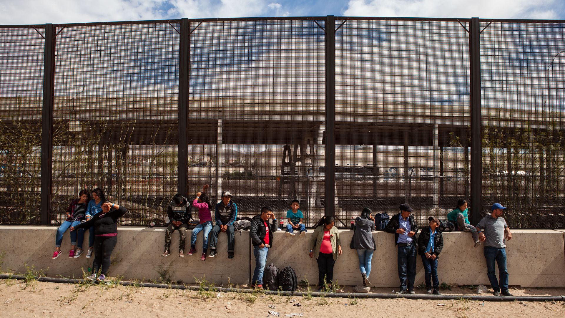 Central American migrants standing in front of a U.S.–Mexico border wall