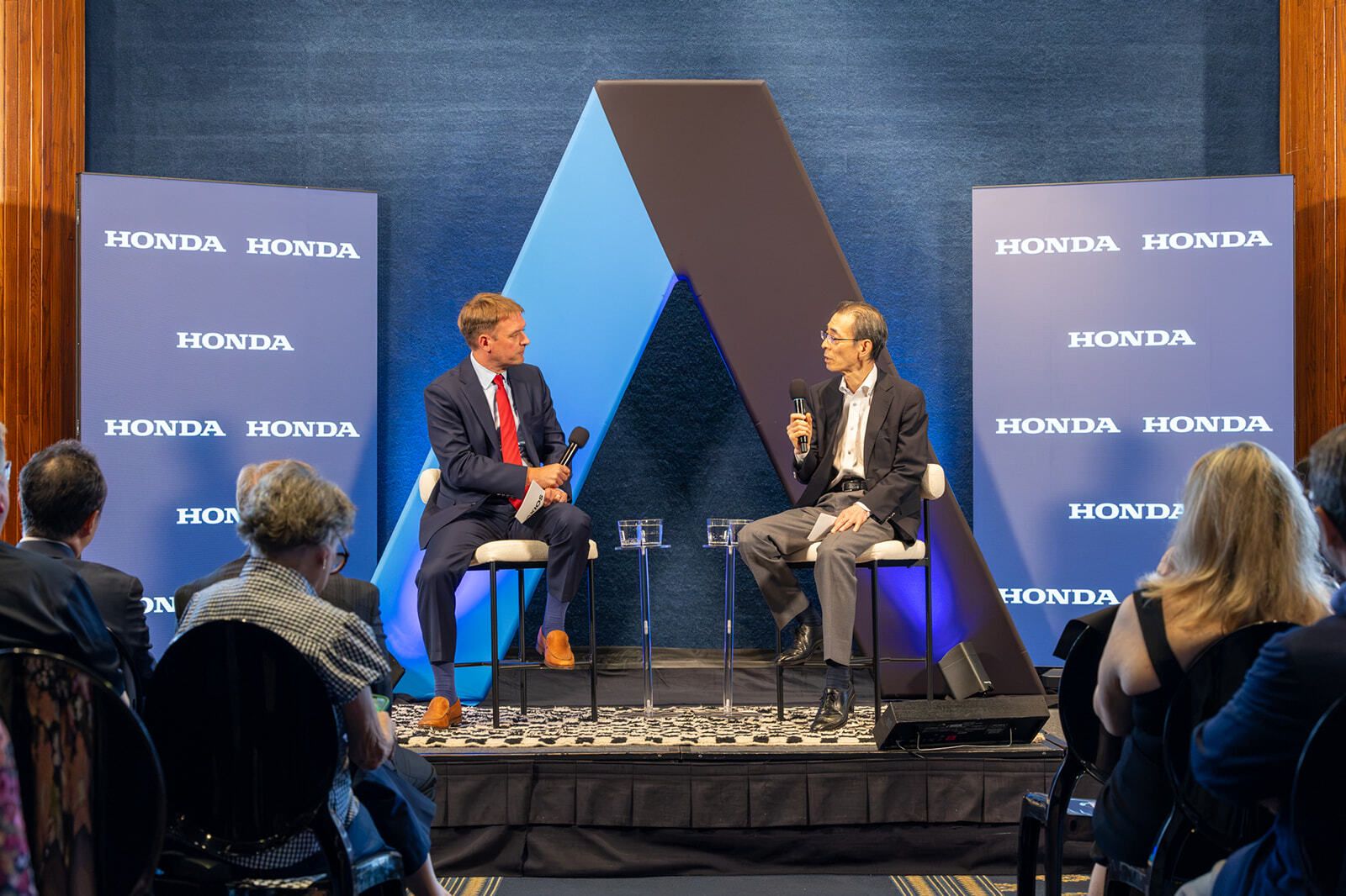 Two men in suits sit on stage with microphones at a Honda event. They chat before a blue and black geometric backdrop, flanked by tall blue HONDA banners; audience seated in front.