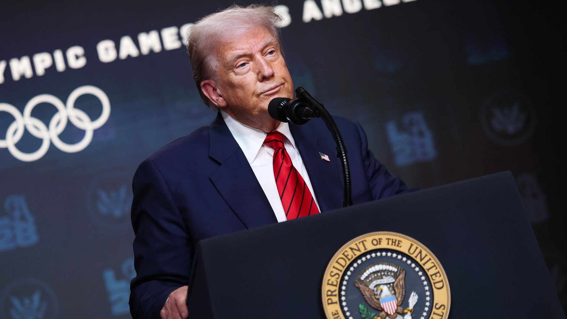 Donald Trump in dark suit, white shirt, red striped tie speaking at podium with the Seal of the President of the United States, Olympic rings and text in background.