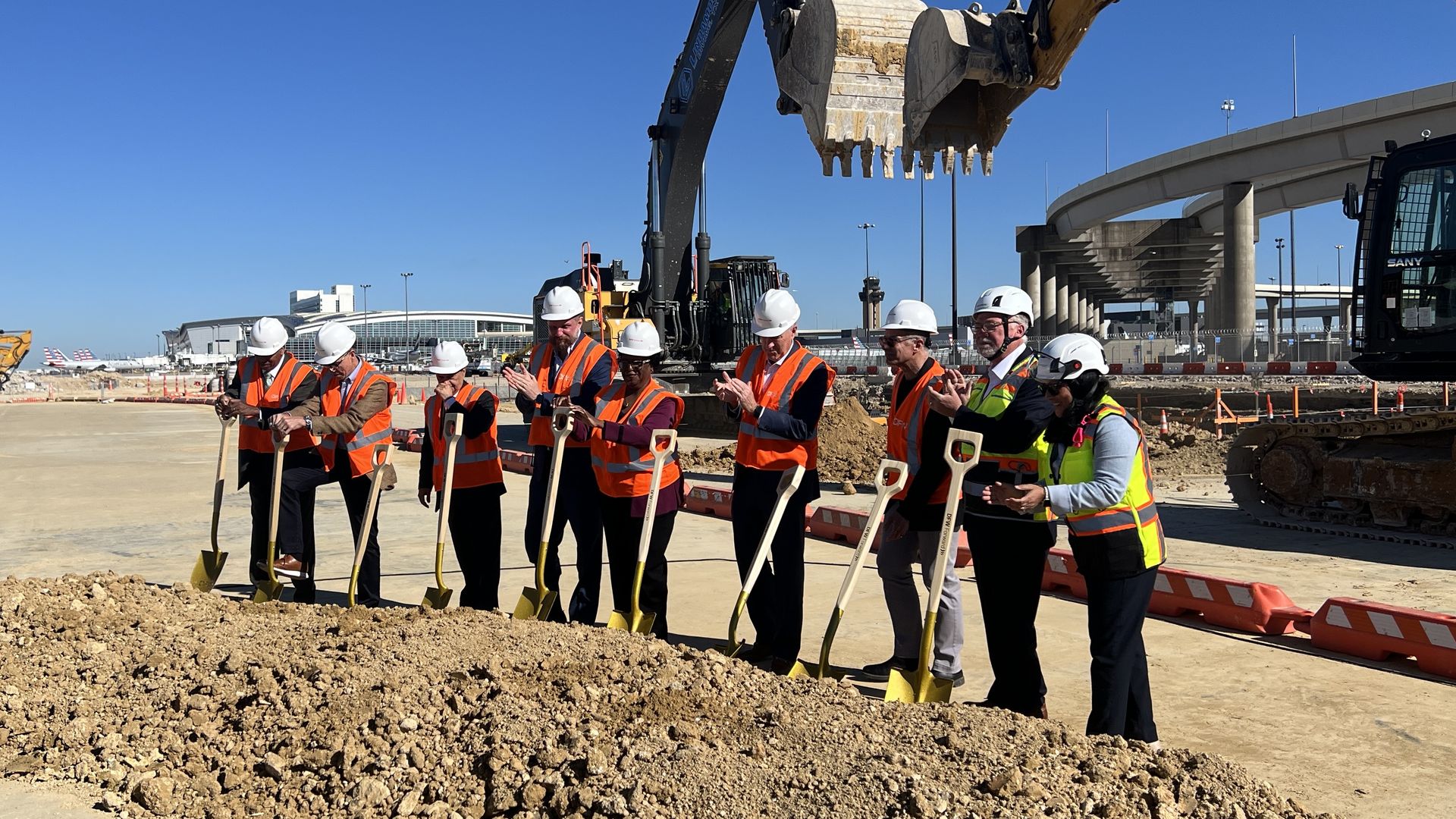 Nine people stand with shovels over a mound of dirt