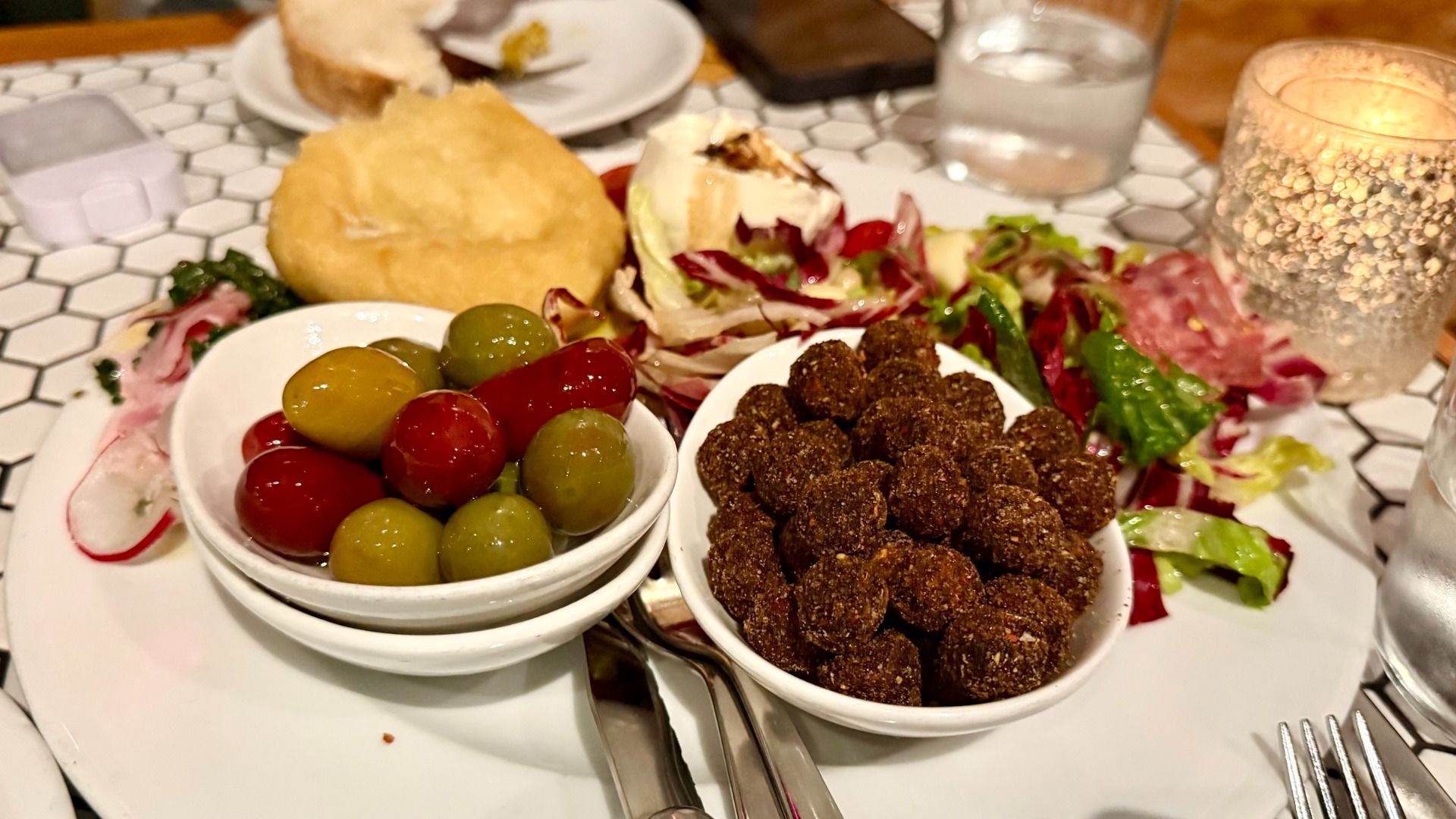 Plate with a bowl of mixed green and red olives, a bowl of dark brown coated nuts, mixed salad greens, sliced meat, a piece of fried bread, and a glass candle on a tiled table.