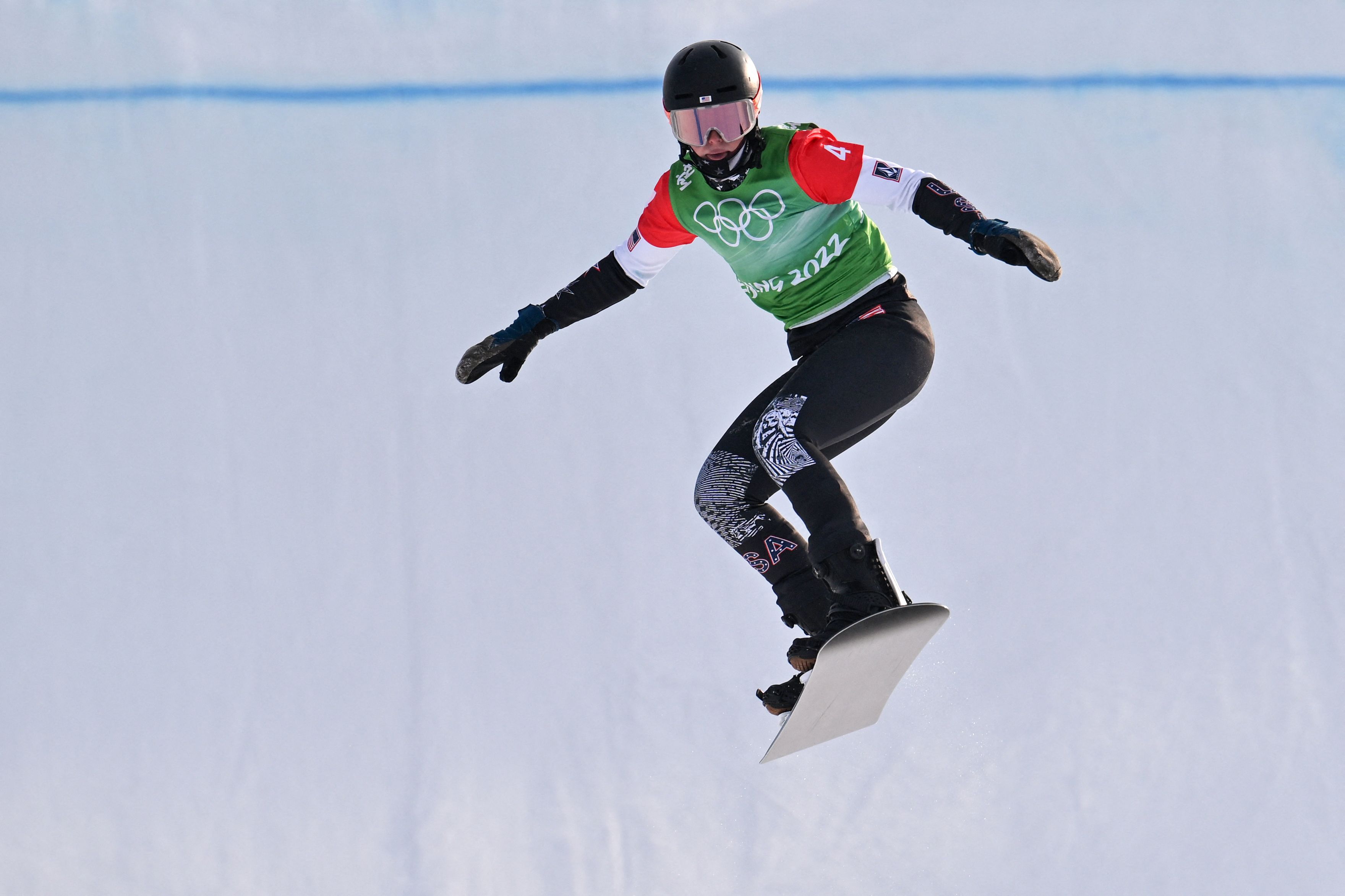 Snowboarder wearing green Beijing 2022 Olympic bib, black helmet, and black pants with patterns, mid-air against snowy background during a jump.