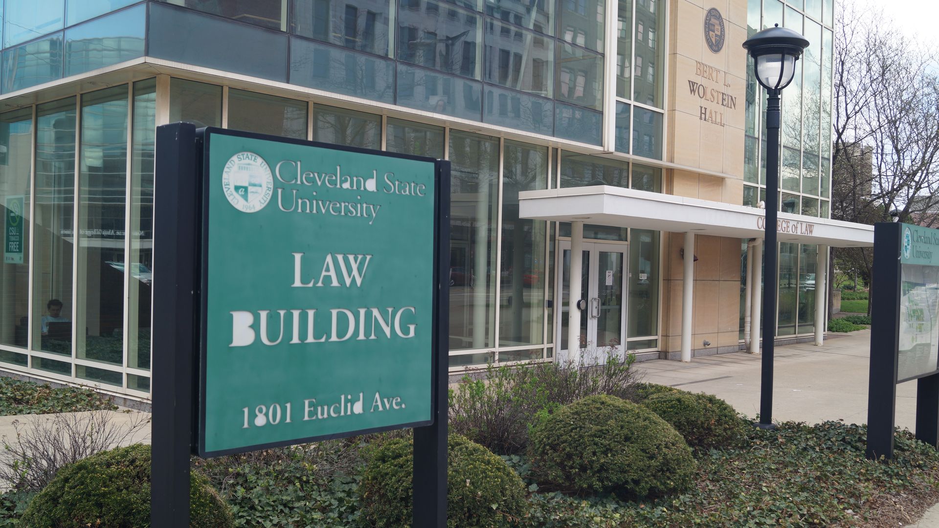 Teal sign for the Cleveland State University Law Building reading 1801 Euclid Ave in front of a glass-walled campus building with a white canopy, lamp post, and trimmed shrubs.