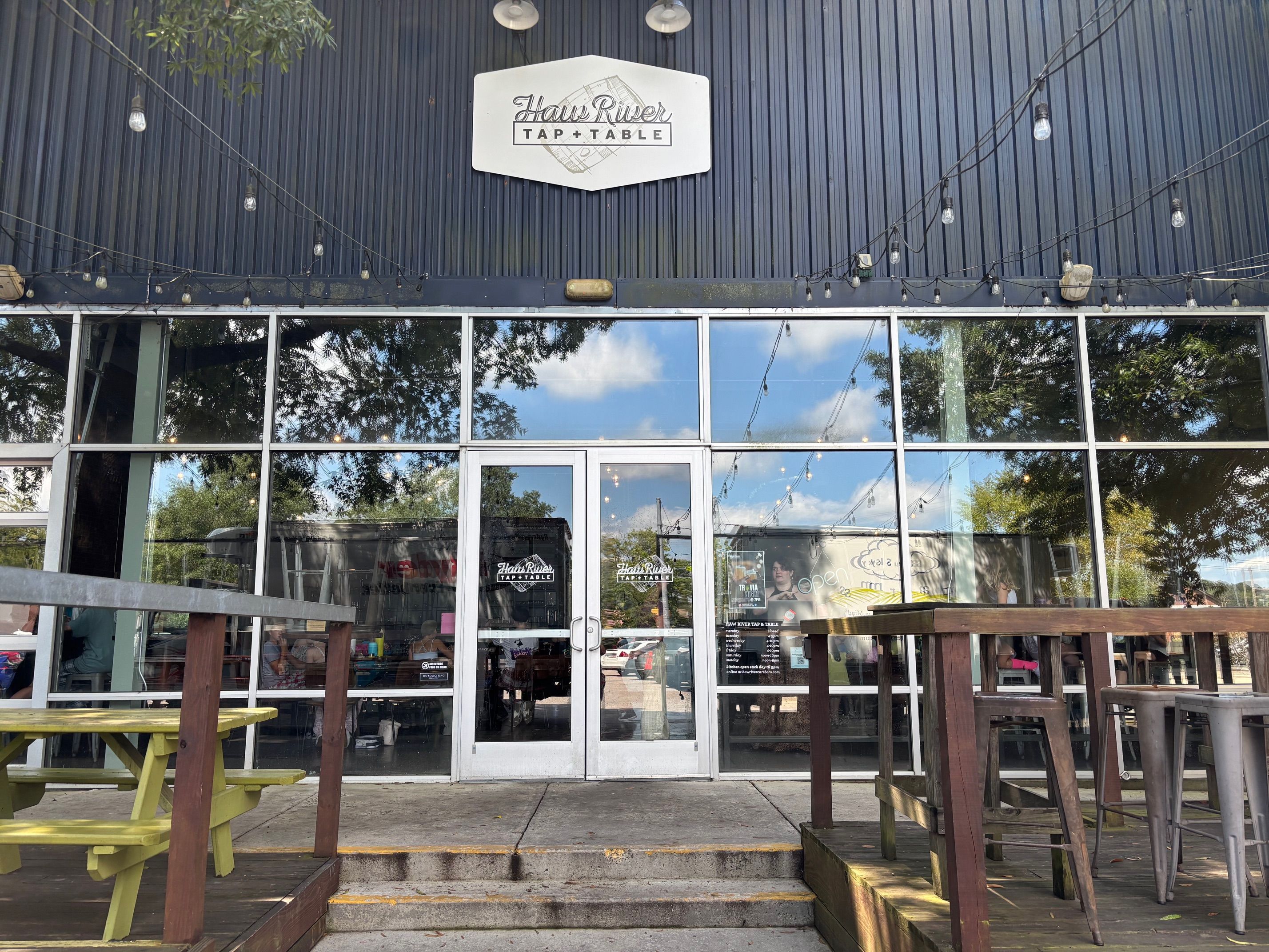 Front entrance of Haw River Tap + Table with glass doors and windows, outdoor seating with yellow picnic table on left and tall wooden tables with metal stools on right, string lights overhead.