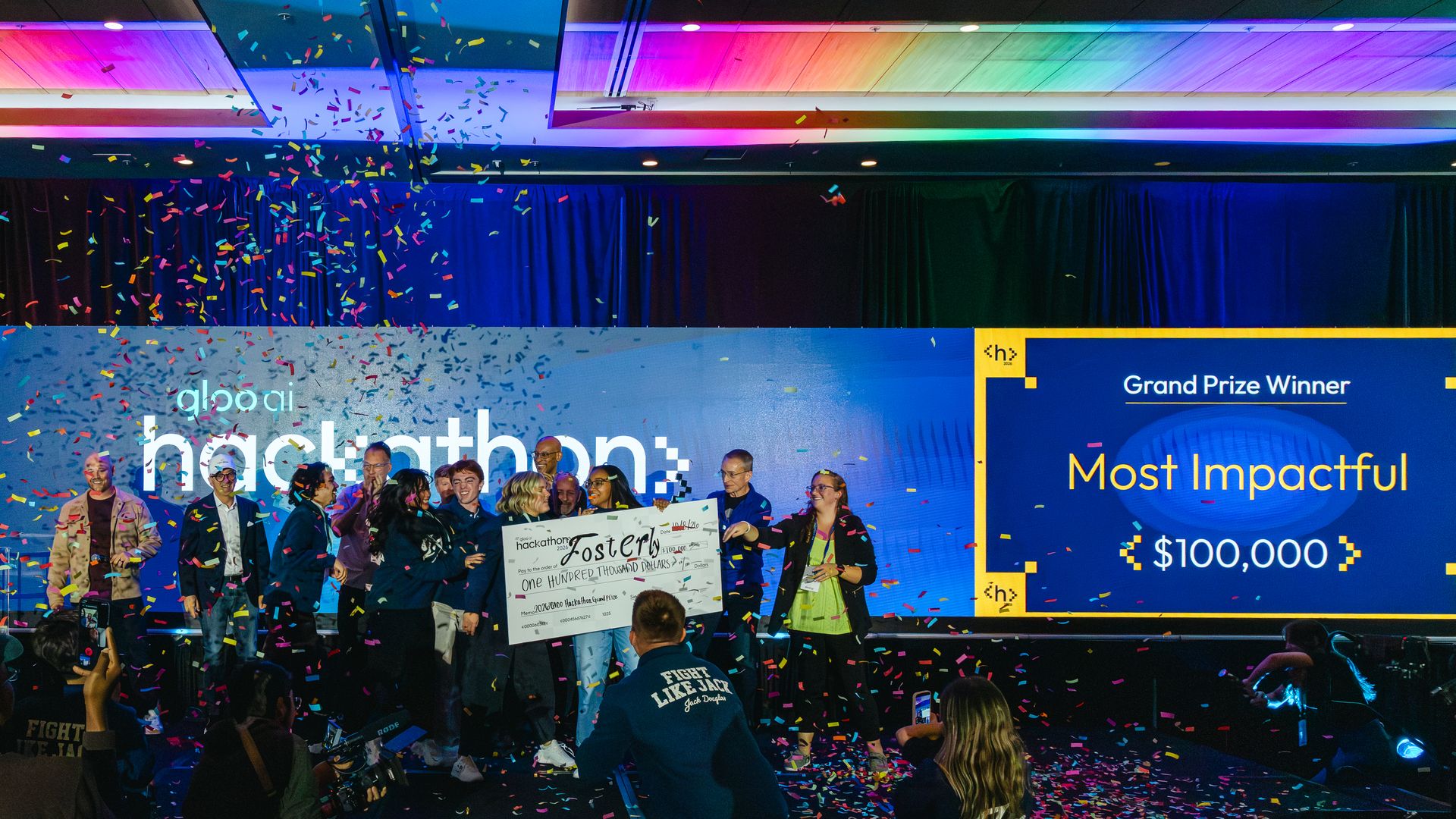 Group of people on stage at a hackathon awards ceremony, holding a large check amid multicolored confetti. Bright blue banners read hackathon and Most Impactful as prizes are awarded.