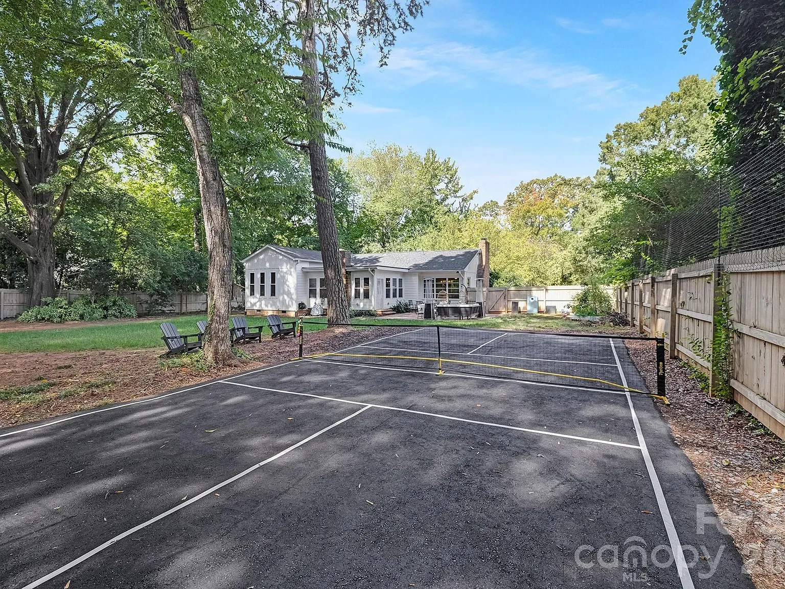 Backyard with a black asphalt tennis court and net in the foreground, a white house with a patio in the center, surrounded by tall green trees and a wooden fence.