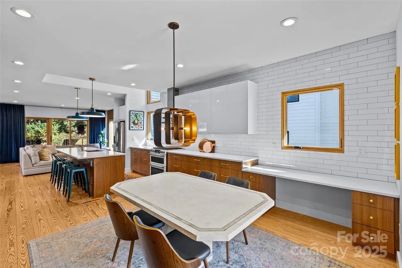 Modern kitchen and dining area with wood floors, white subway tile walls, wood and white cabinetry, marble table with dark chairs, blue pendant lights, and large windows.