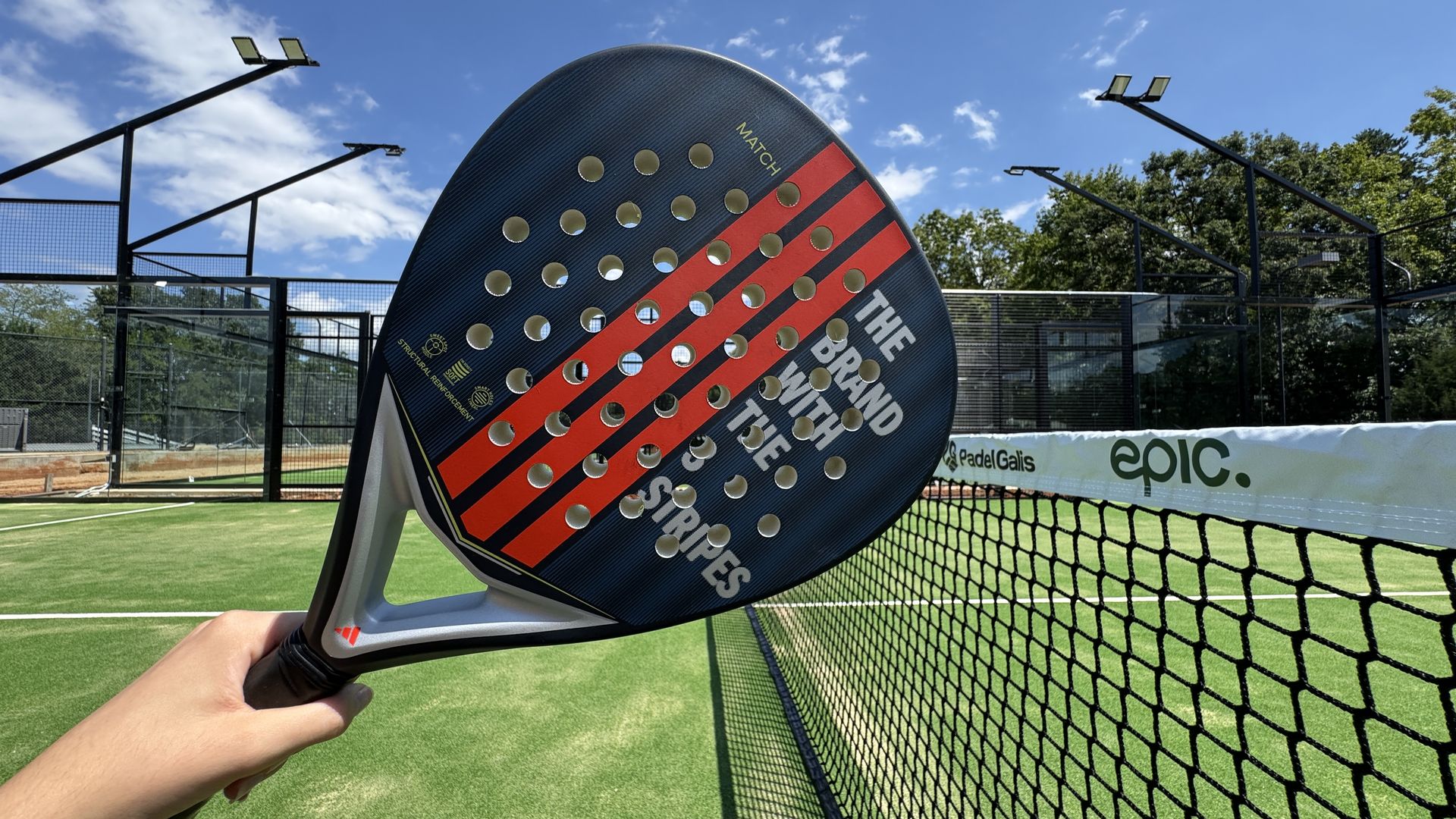 Hand holding a black and red padel racket with text, over a green padel court with a net labeled "epic." and "PadelGalis" under a blue sky with scattered clouds.
