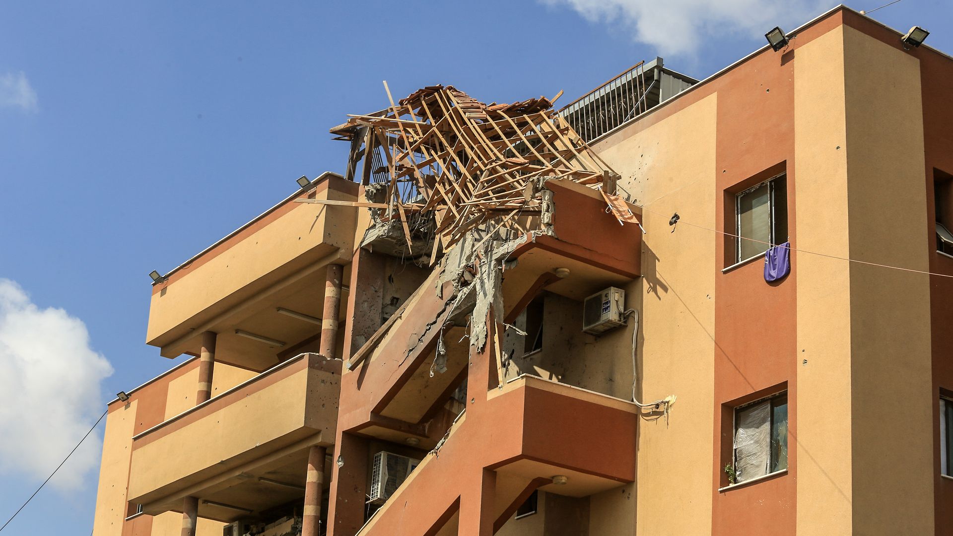 Damaged orange and beige building with destroyed staircase roof and rubble hanging from the edge against a clear blue sky with some clouds.