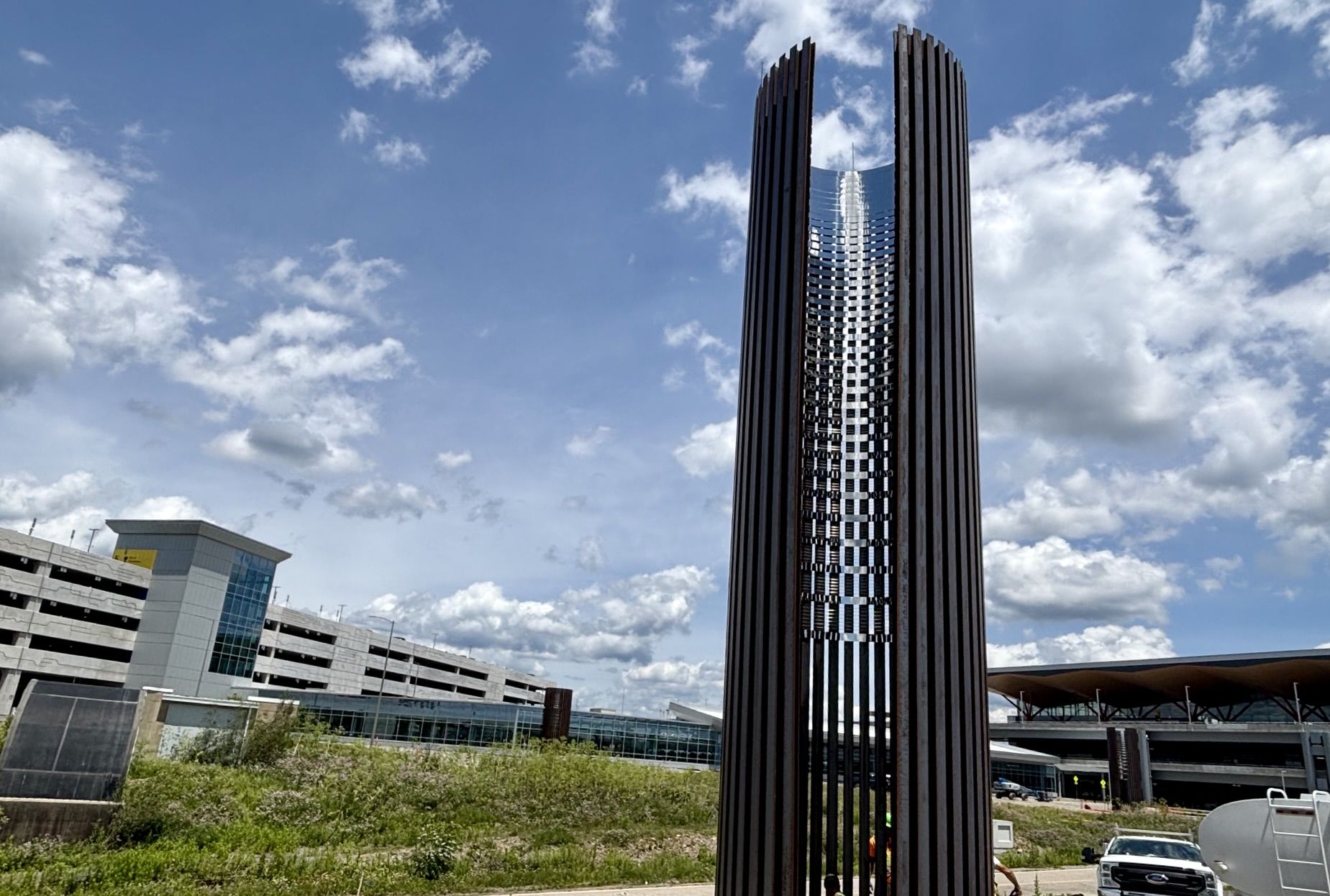 Tall cylindrical sculpture with vertical dark slats around a reflective core, set against a blue sky with scattered clouds; a modern parking structure and greenery lie in the background.