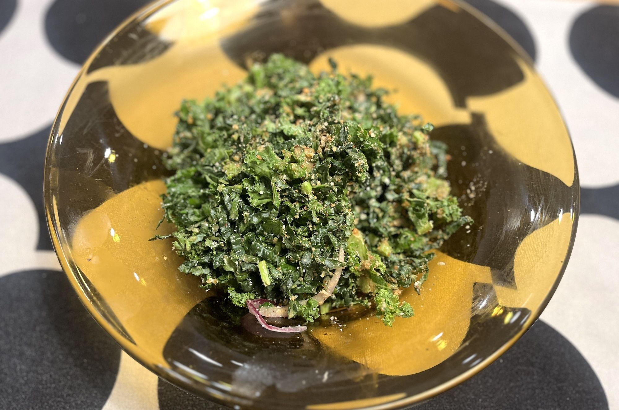 Close-up of chopped green kale with breadcrumb crumbs on top, served in a yellow-brown patterned glass bowl on a black-and-white patterned table.