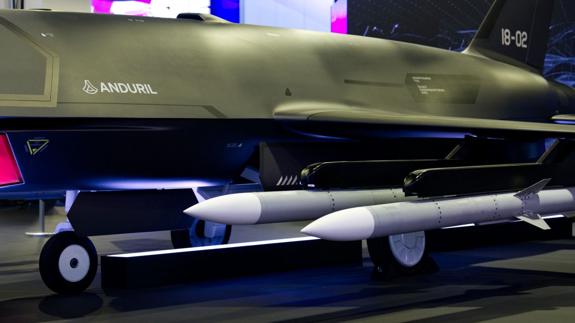 Side view of a large military drone on display, with the Anduril logo on the fuselage, tail number 18-02, and white missiles mounted underneath.