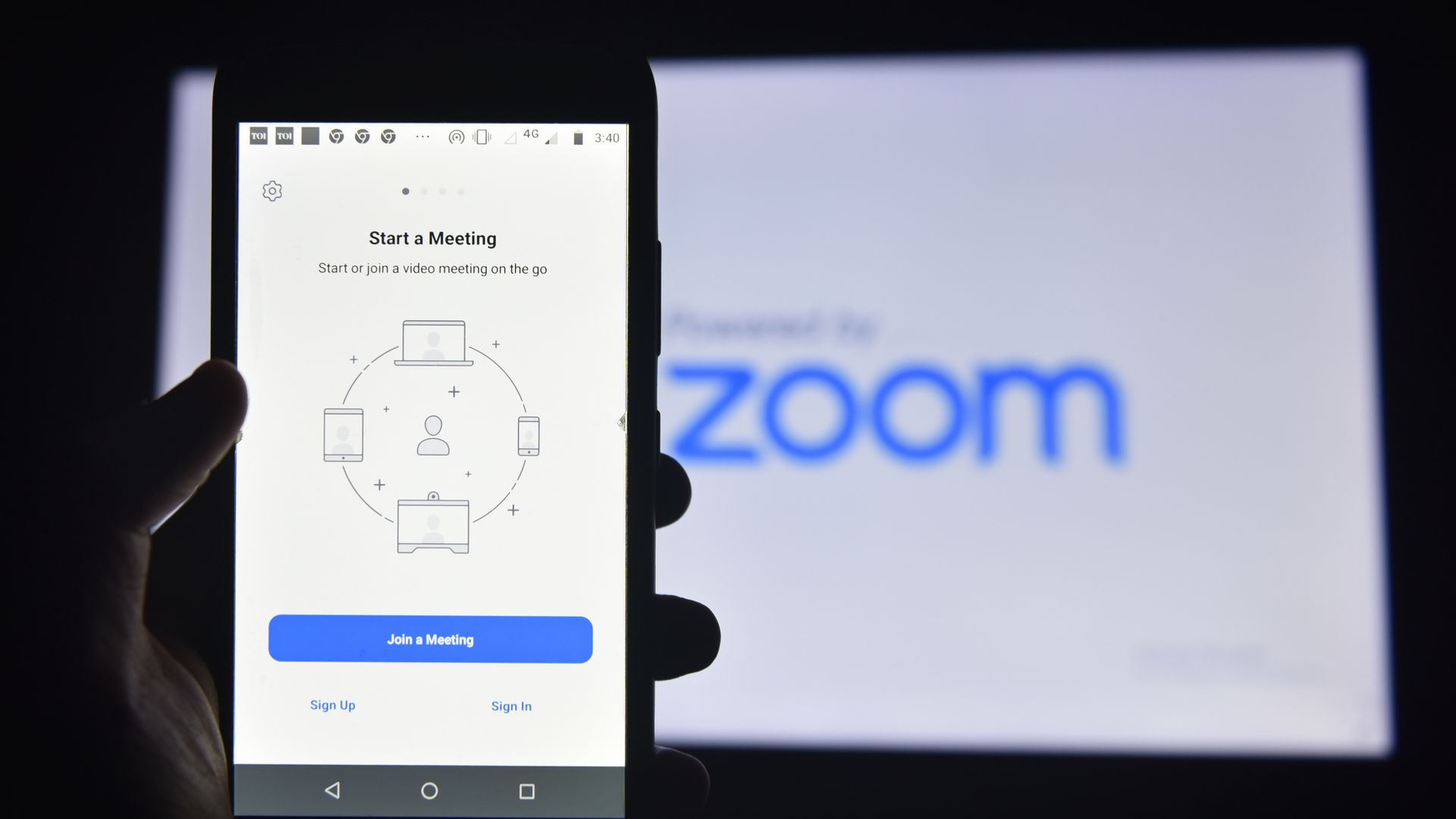 A photo of a smartphone displaying a Zoom meeting screen, with a larger screen displaying the Zoom logo in the background.