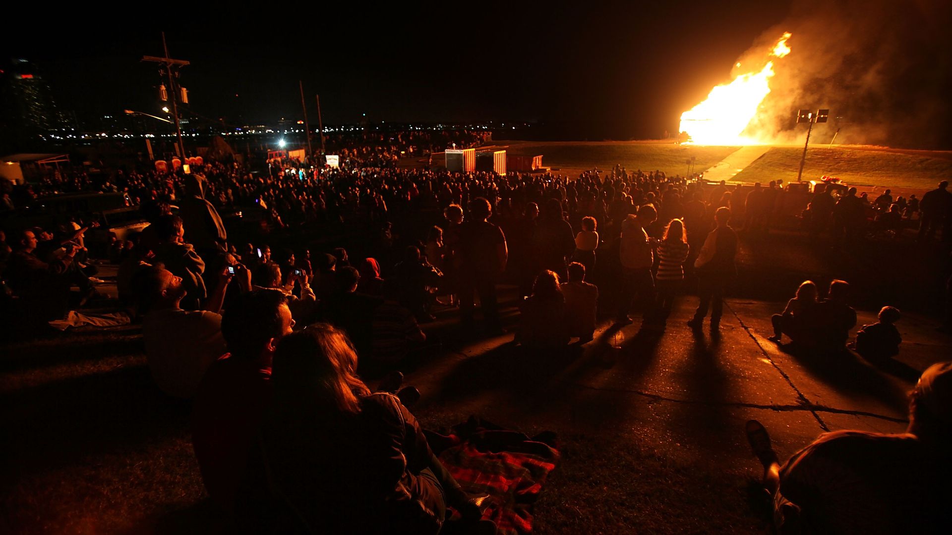 A crowd of people watch a bonfire from a levee. They're all lit by the glow of the burning fire.
