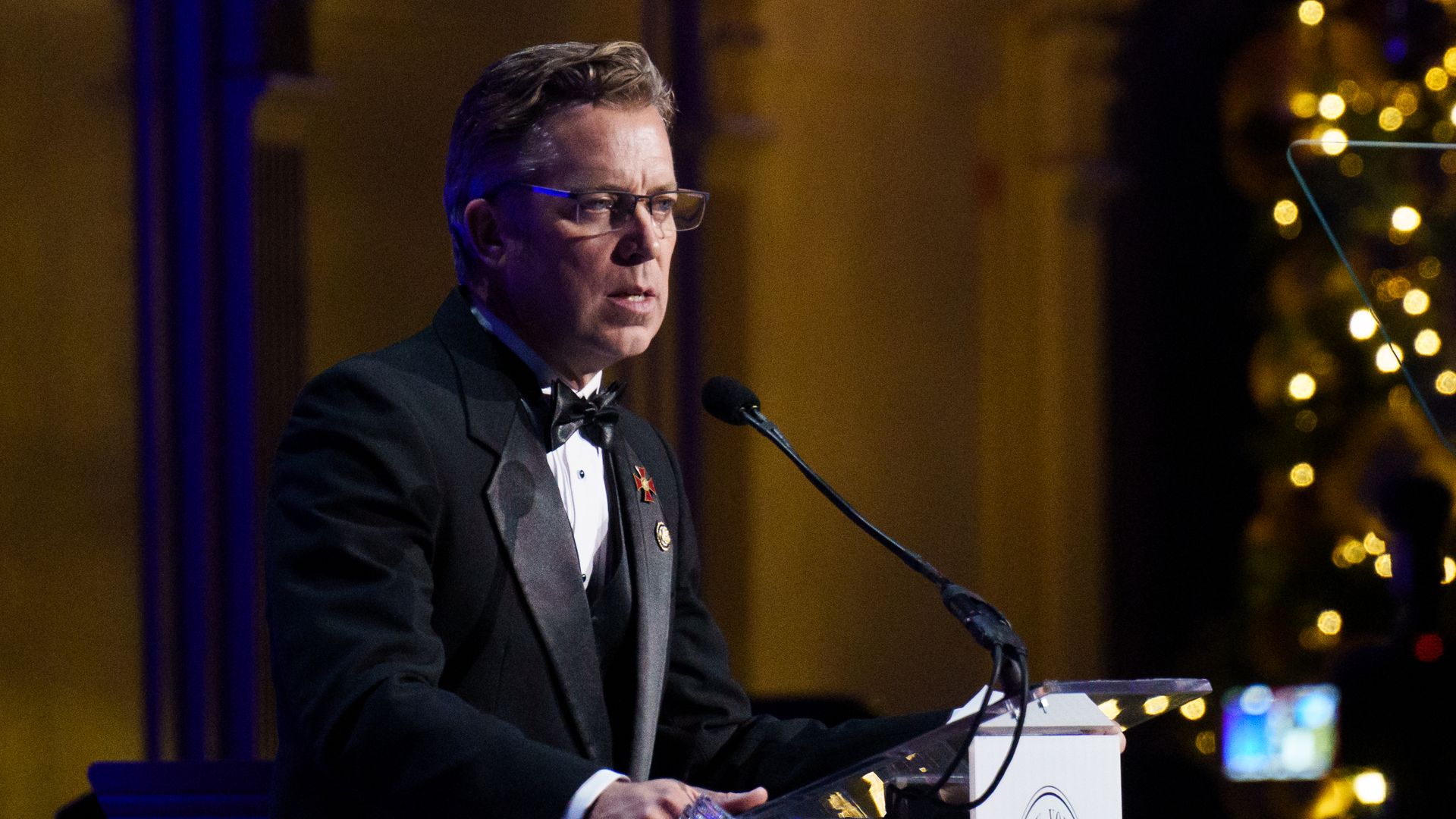 A man in a black tuxedo and bow tie, wearing glasses, speaks into a microphone at a podium with an emblem in a dim, warm-lit venue, holding a water bottle as gold bokeh lights glow behind him.