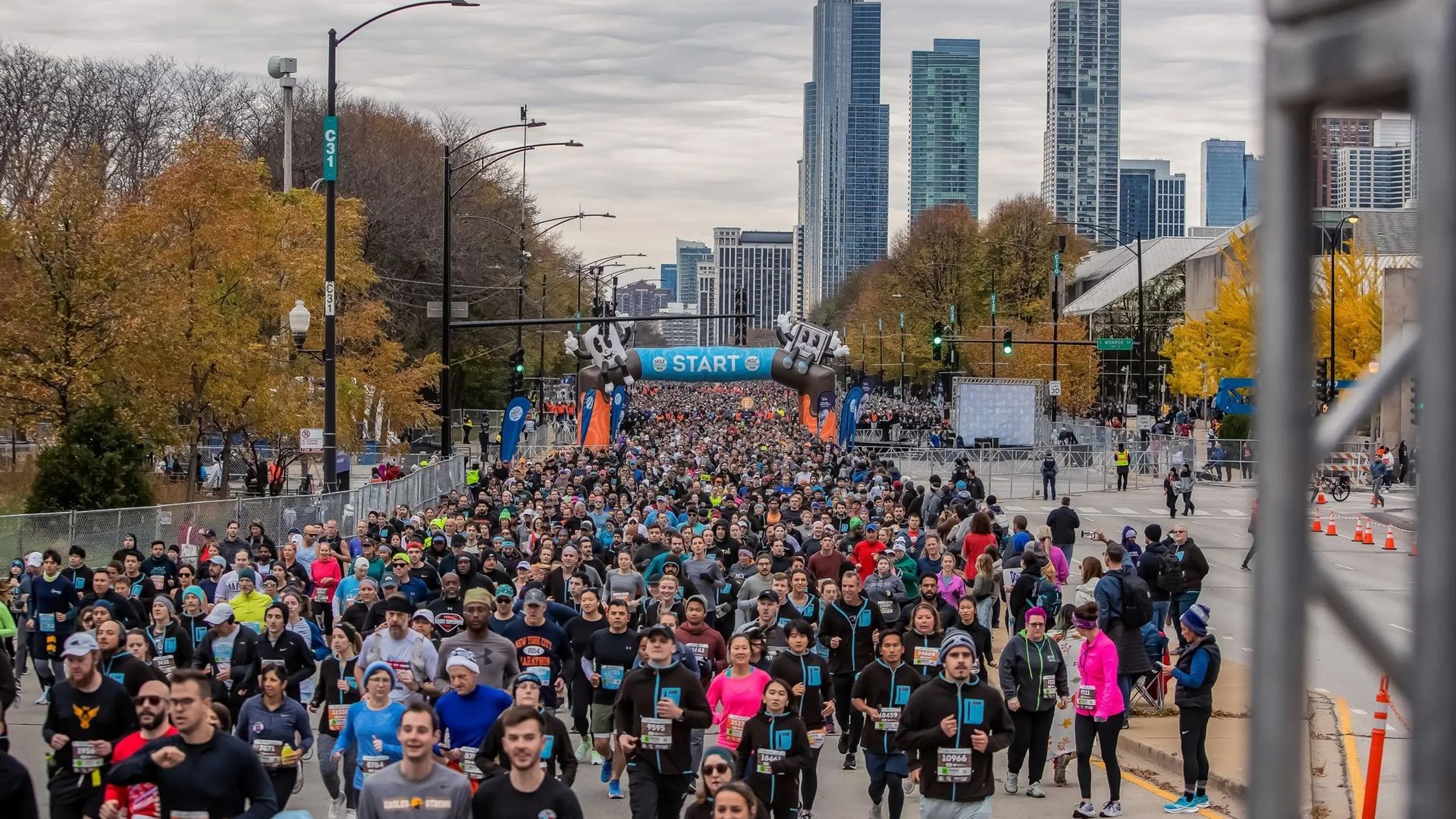 A large crowd of runners gathers at a race start line with a blue "START" banner, autumn trees, and a city skyline with tall buildings under a cloudy sky.