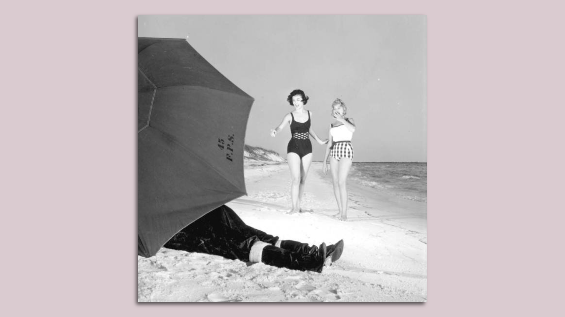 Two women in vintage bathing suits on the beach pointing at an umbrella with just what looks like Santa Claus' boots sticking out from underneath it.