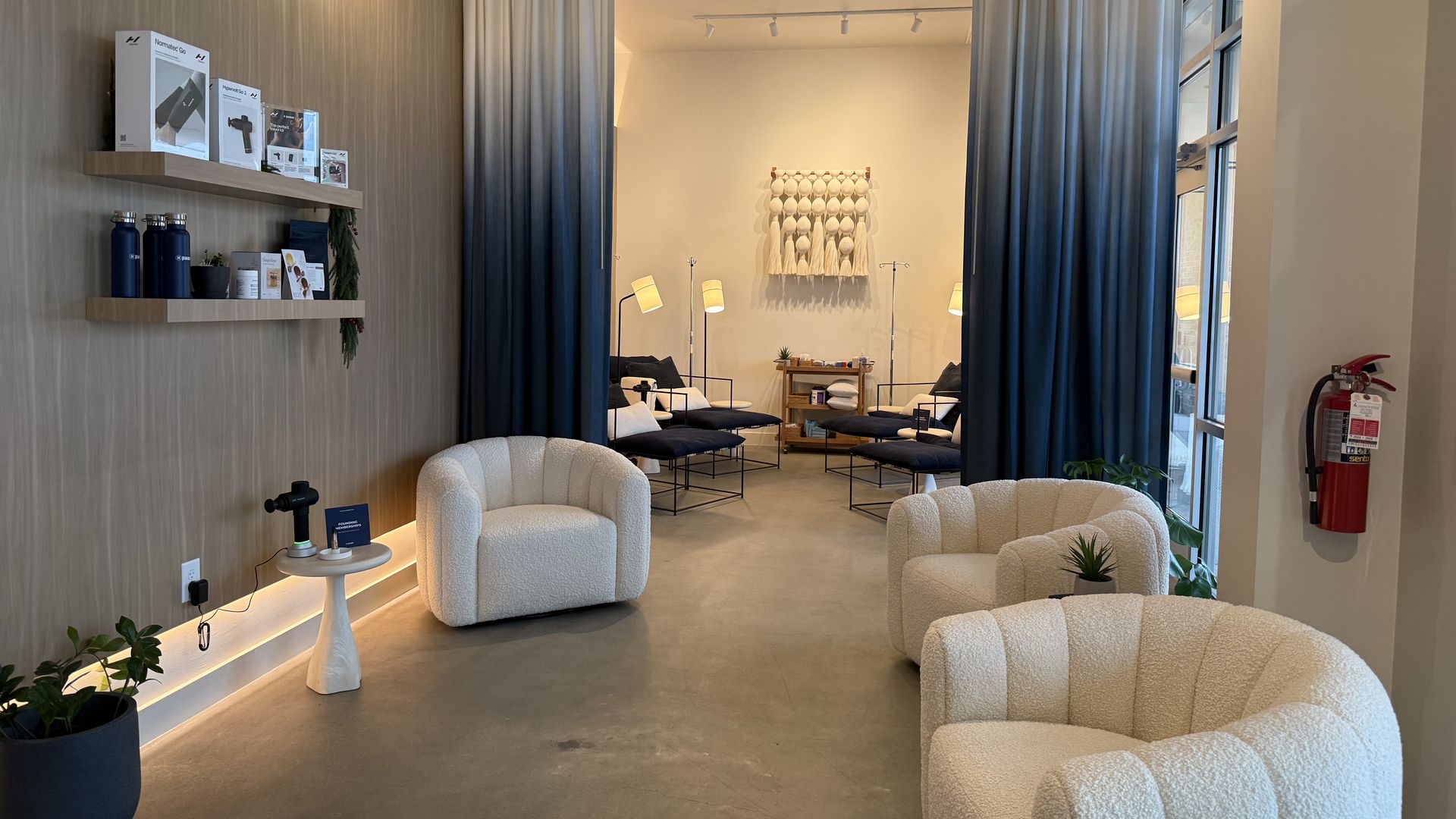 Bright lounge with white curved armchairs, blue gradient curtains, modern floor lamps, wood shelves displaying products on the left wall, a large window glass door, and a fire extinguisher.
