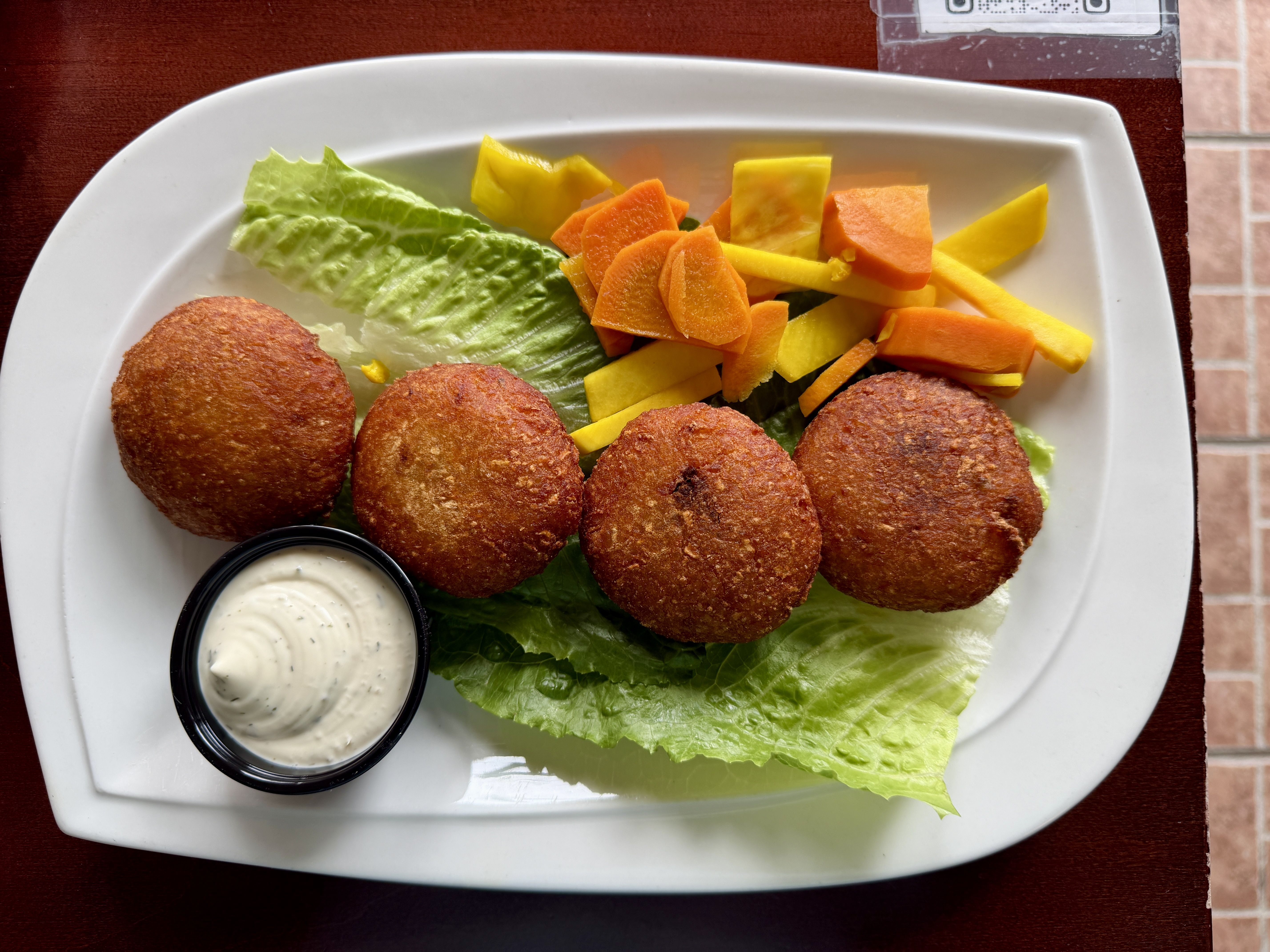 Four golden-brown croquettes arranged on green lettuce on a white plate, with a small container of creamy dip and colorful carrot and yellow mango sticks on the side.