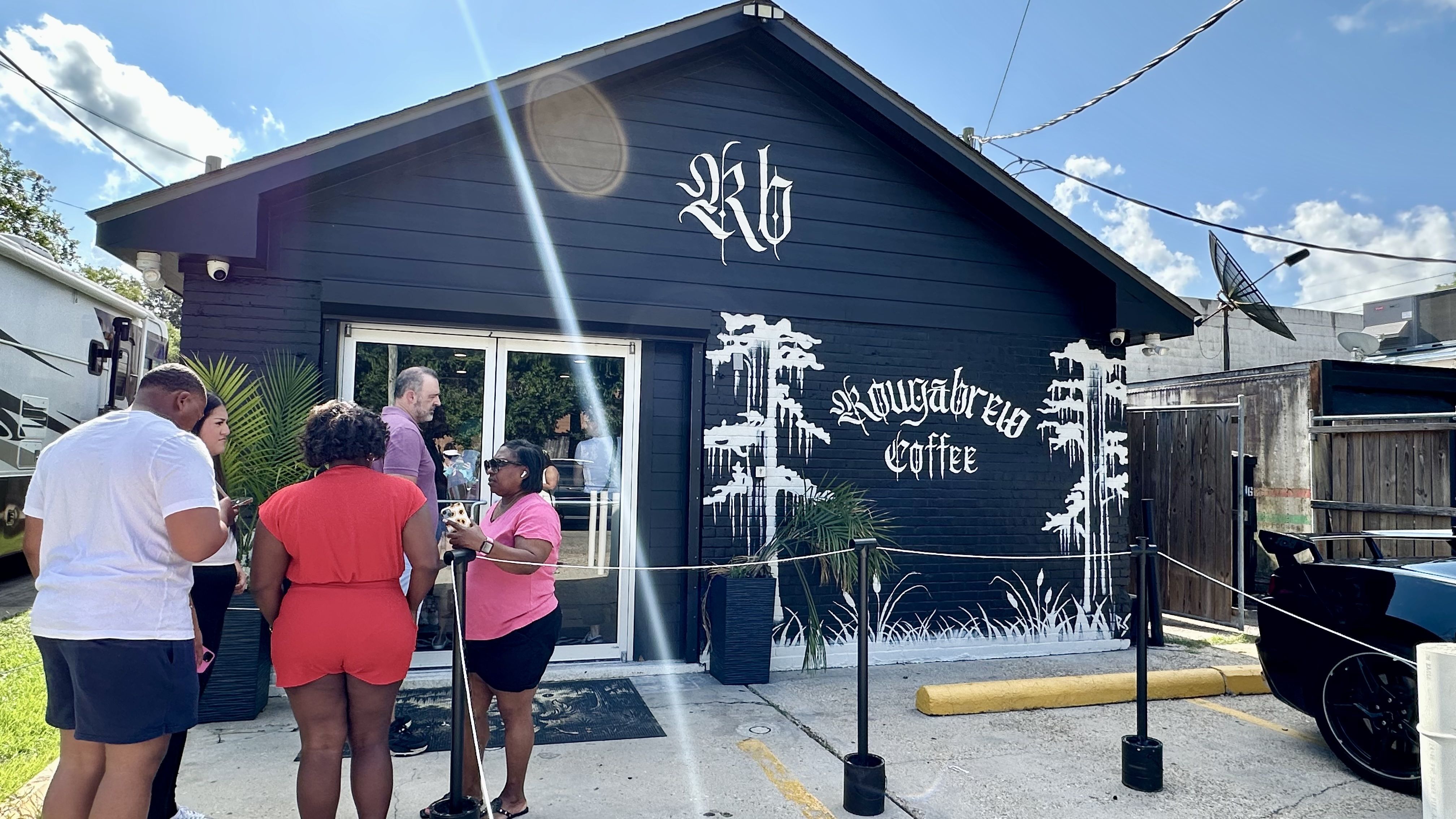 People stand outside a dark blue coffee shop with ornate white lettering on the wall that says Coffee; sunny day, glass entrance, a palm plant, and a satellite dish on the side.