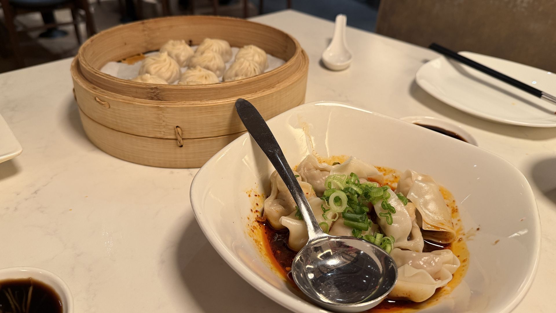 Bamboo steamer with dumplings on a white table, beside a bowl of dumplings in orange sauce and green onions. A spoon rests in the bowl; chopsticks lie on a plate, with a blurred restaurant background.