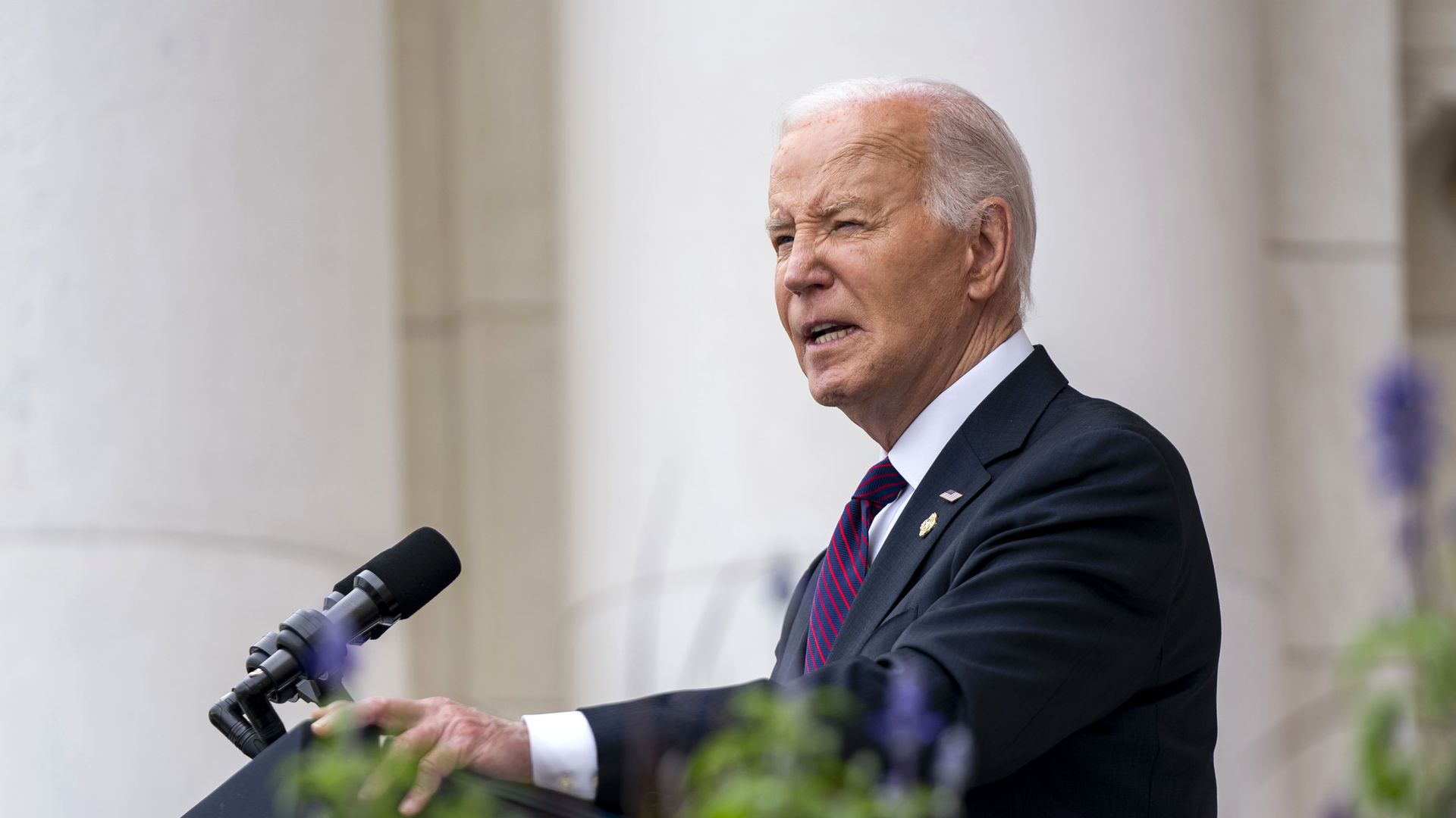 President Biden, wearing a blue suit, white shirt and red and blue tie, speaks at a microphone.