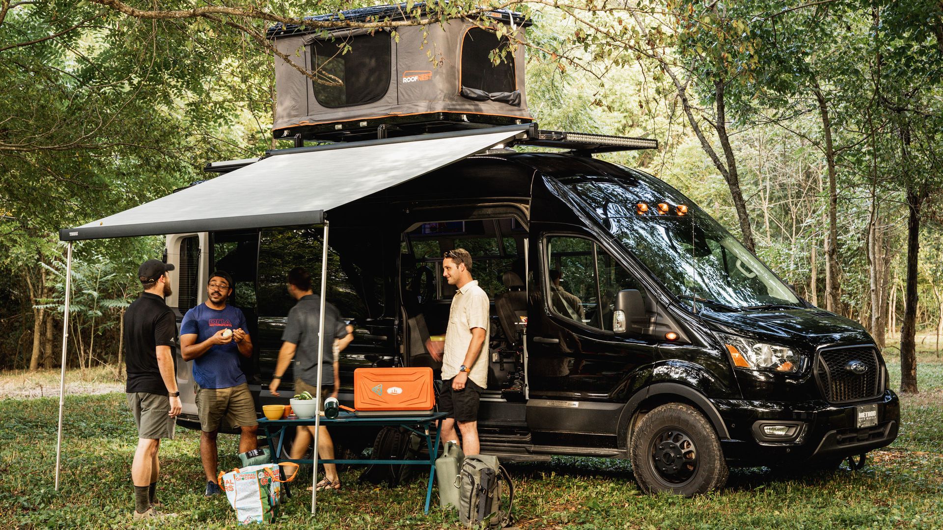 Three men stand around a camper at a campsite in the woods.