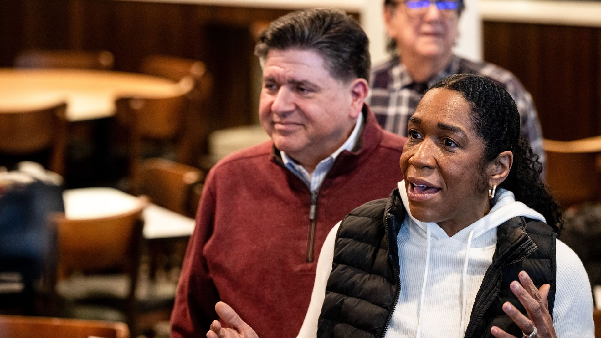 Woman in a white hoodie and black puffer vest speaks at a press conference, gesturing with her hands. A man in a maroon sweater stands behind her as microphones—including FOX 32 Chicago—line the foreground.
