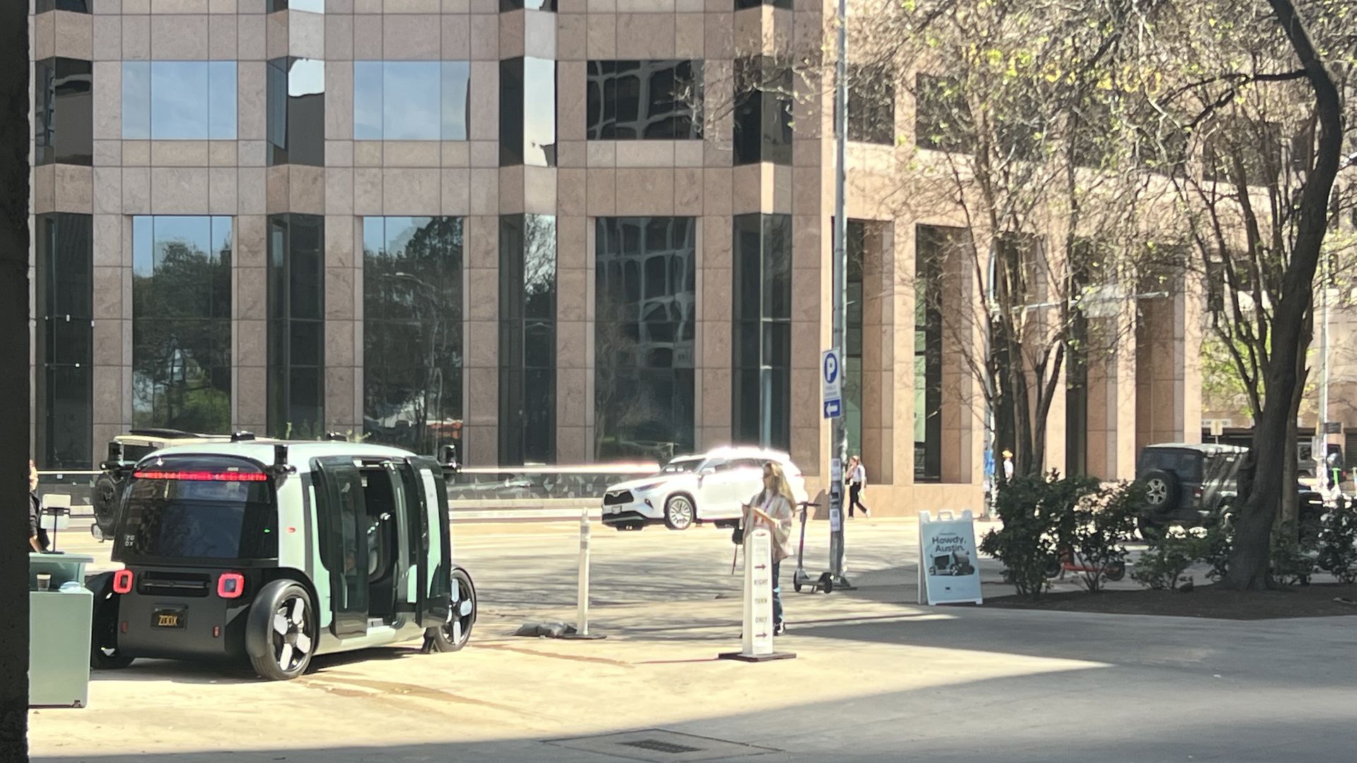 Wide street view of a beige, glass-windowed office building with tan panels. Right side shows leafless trees; foreground has parked cars, a bus, and a person walking along the sidewalk.