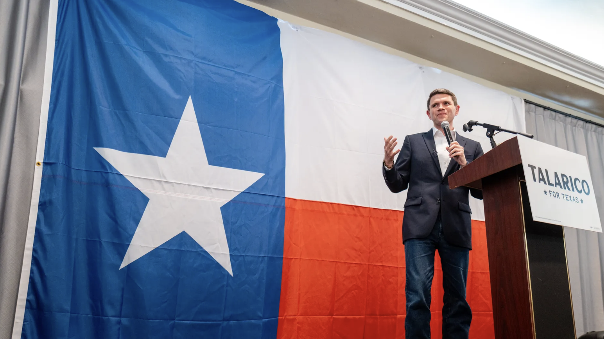 Male speaker in a navy blazer and jeans stands at a wooden podium with a microphone, gesturing. Behind him is a large Texas flag backdrop with blue, white, and red panels; the podium reads "TALARICO FOR TEXAS".