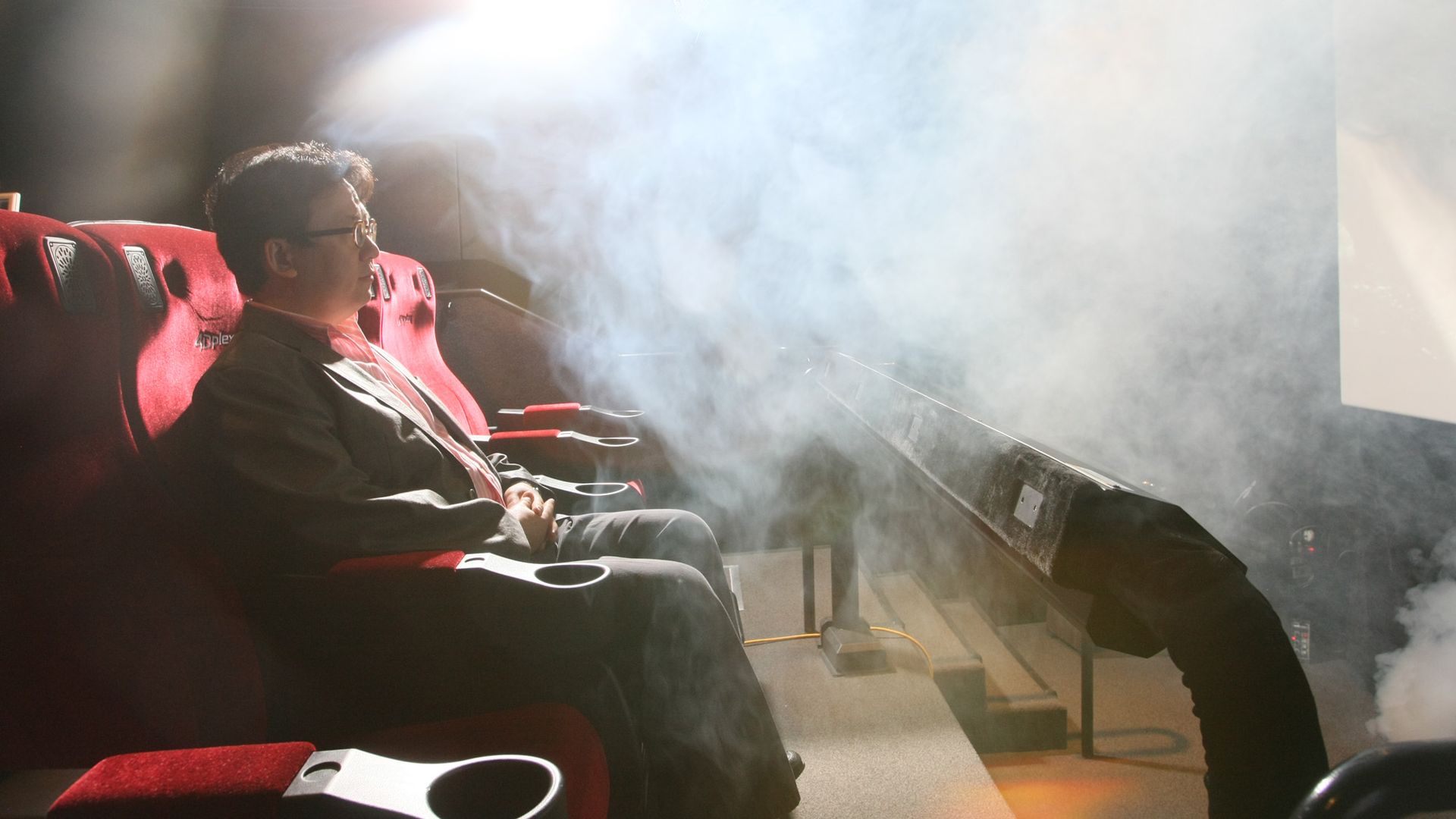A man in a suit and glasses sits in a red theater seat as thick theater fog and a bright stage light fill a dim auditorium.