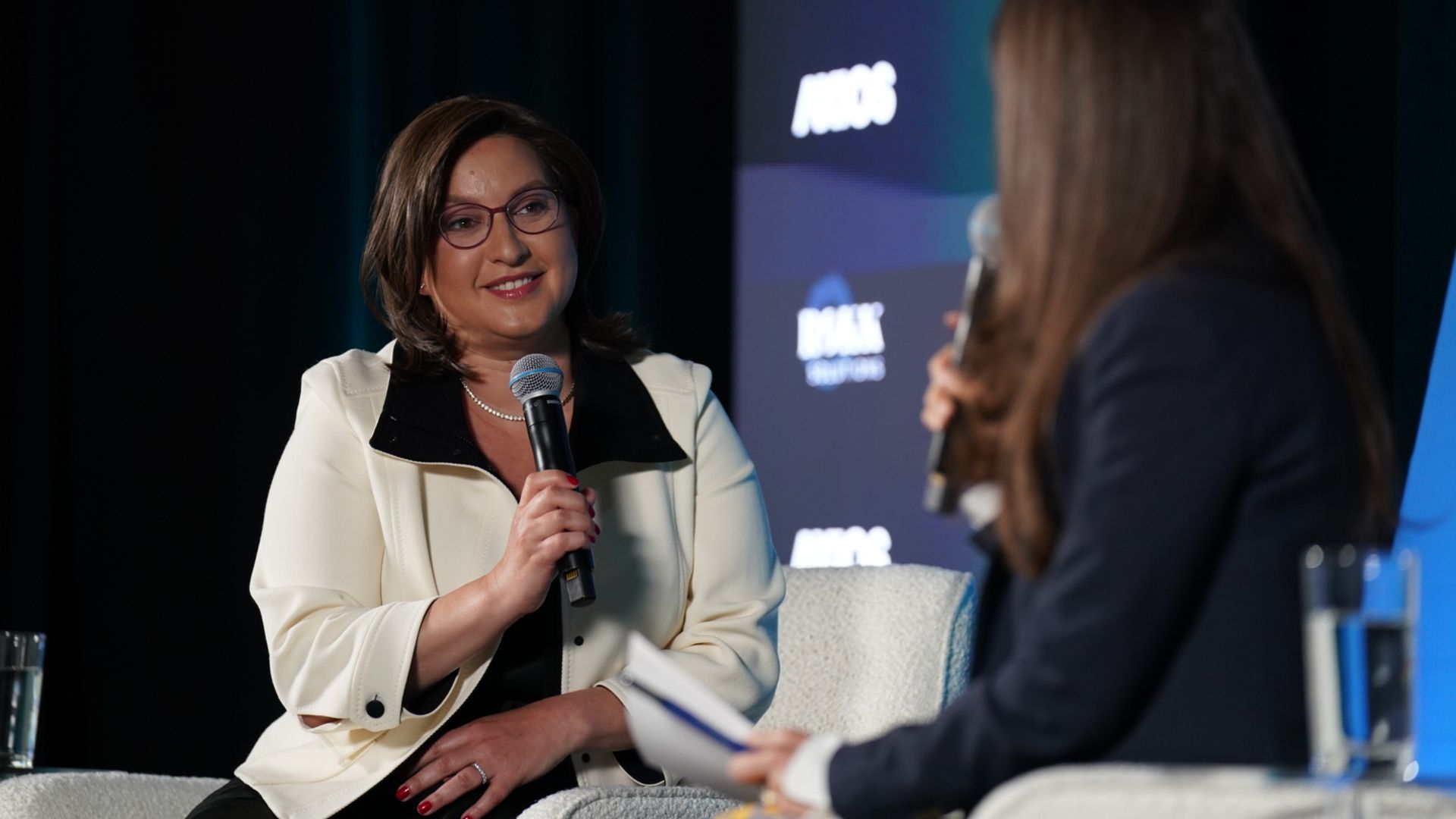 Two women on a stage interview. Left: Rebecca Kutler in a cream blazer with black lapels, pearl necklace and red glasses, holding a microphone. Right: Sara Fischer in dark clothing; backdrop with logos.